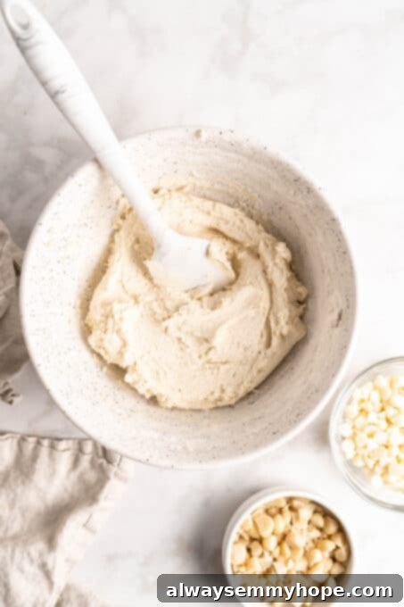 Wet ingredients mixed for vegan white chocolate macadamia nut cookies. Overhead view of a bowl of creamy cookie dough, with small bowls of precisely chopped macadamia nuts and sweet white chocolate chips ready to be incorporated.