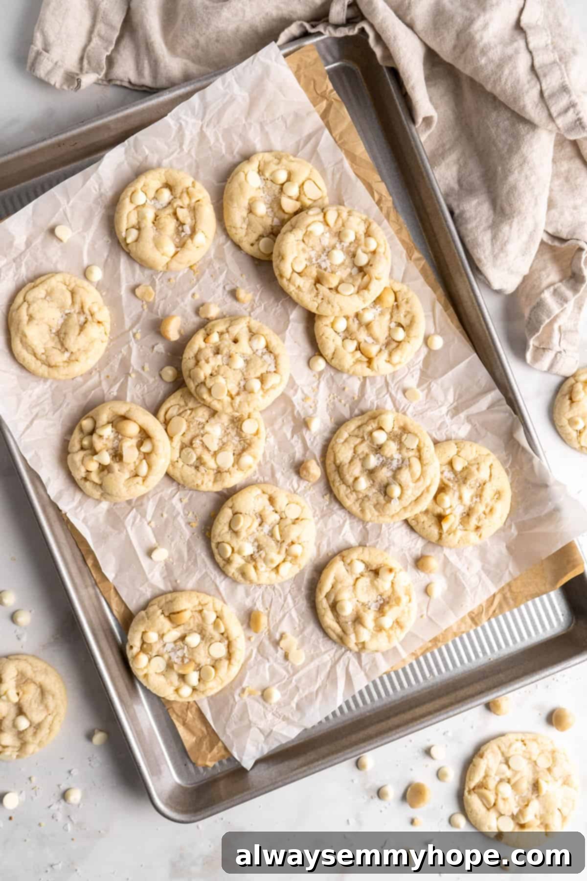 Assorted vegan white chocolate macadamia nut cookies cooling. Overhead view of an assortment of baked vegan white chocolate macadamia nut cookies, some with cracks, cooling on a parchment-lined baking sheet.