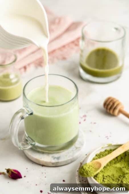 Frothed plant-based milk being poured gracefully into a clear glass mug containing a bright green matcha base, creating a beautiful layered latte.