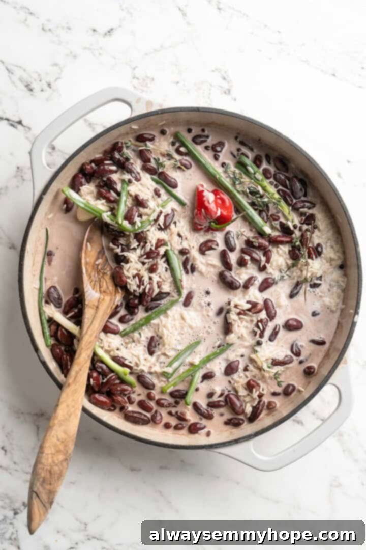 This authentic Jamaican rice and peas recipe is an easy side dish with lots of island vibes! It's packed with flavour and so simple to make. Coconut milk, uncooked rice, and various aromatics like scallions and thyme being added to a pan containing cooked kidney beans, showing the combination of ingredients.