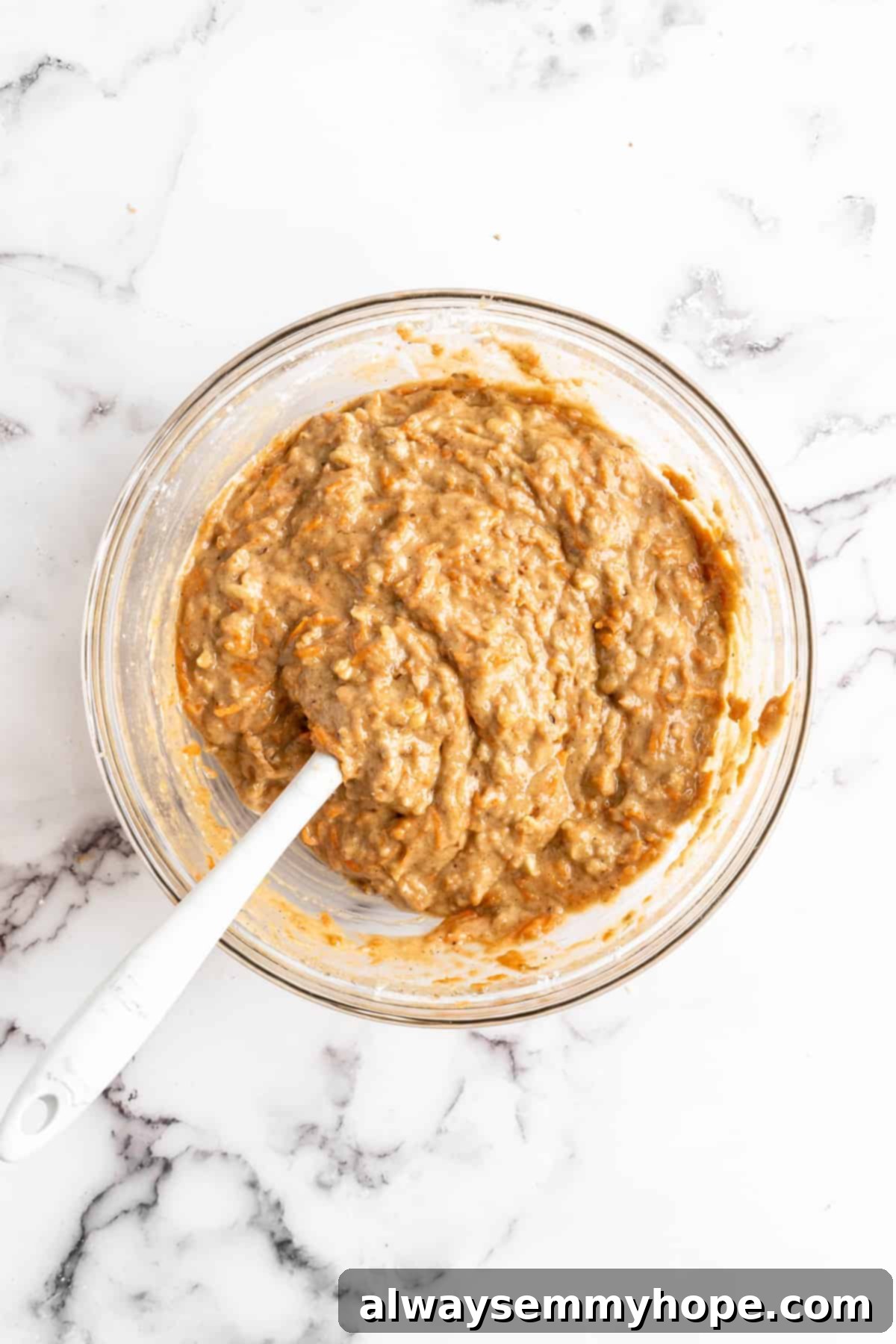Overhead view of carrot cupcake batter in glass mixing bowl with spatula