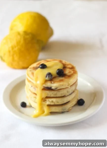 Blueberry Lemon Pancakes with Lemon Curd will brighten any day! Fresh lemon juice, real lemon curd, and blueberries make them extra special. Stack of fluffy blueberry lemon pancakes with a generous dollop of homemade lemon curd and fresh blueberries on a white plate, ready to be enjoyed.