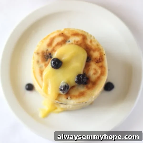 Blueberry Lemon Pancakes with Lemon Curd will brighten any day! Fresh lemon juice, real lemon curd, and blueberries make them extra special. Top down view of a stack of golden brown blueberry lemon pancakes, generously drizzled with glossy lemon curd and garnished with fresh blueberries, on a pristine white plate.