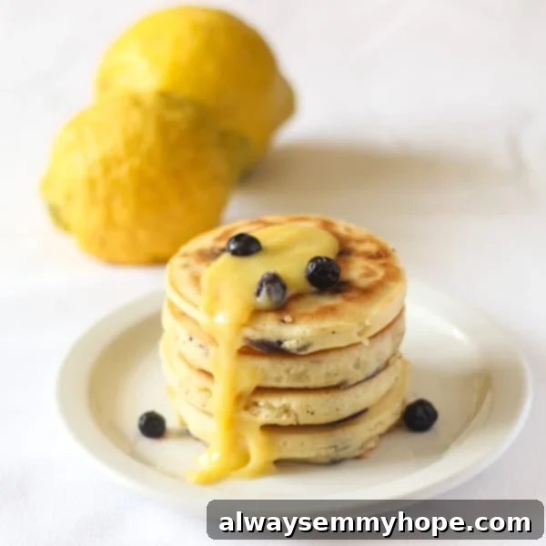 Blueberry Lemon Pancakes with Lemon Curd will brighten any day! Fresh lemon juice, real lemon curd, and blueberries make them extra special. A beautifully arranged plate of blueberry lemon pancakes adorned with fresh lemon slices and juicy blueberries, set against a rustic background with whole lemons, highlighting the freshness of ingredients.