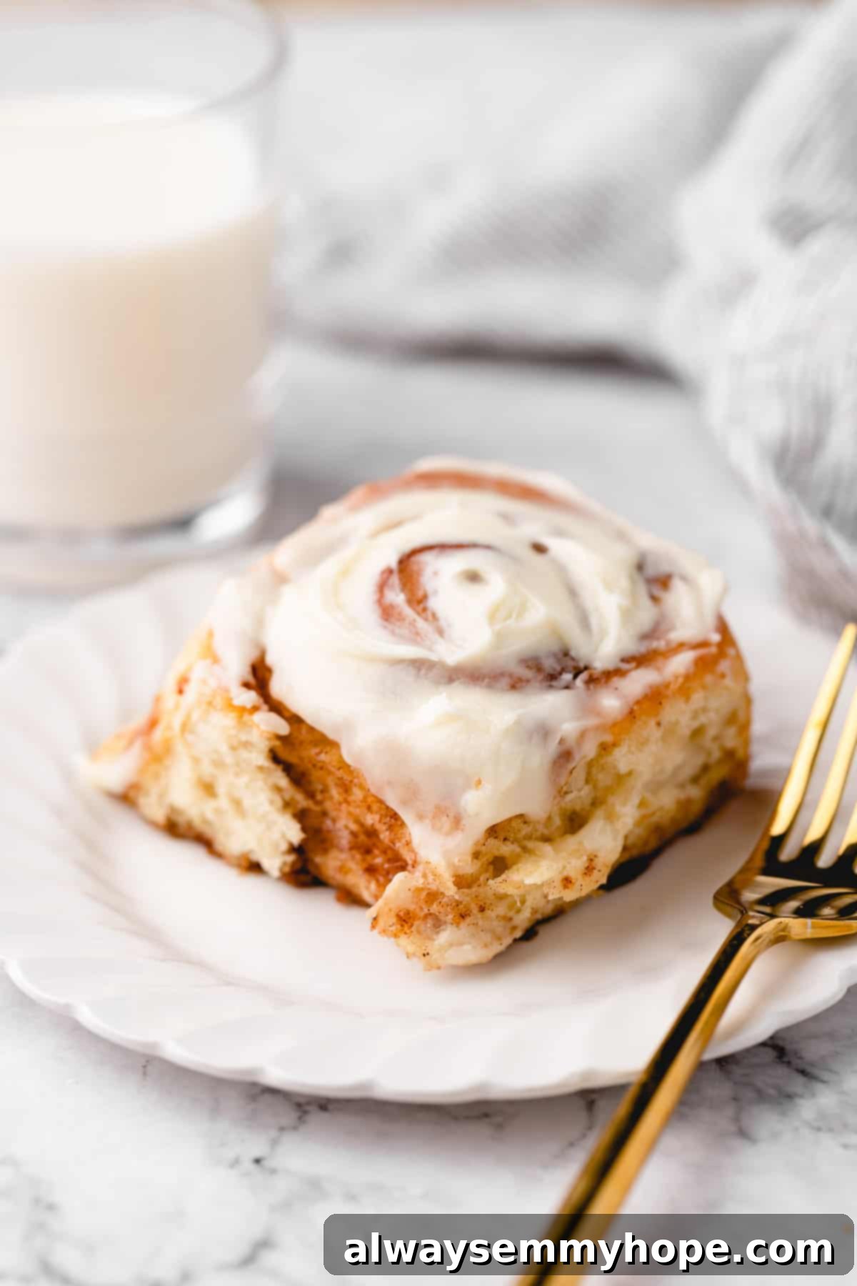 A single perfectly frosted vegan cinnamon roll on a white plate with a gold fork, a glass of plant milk in the background.