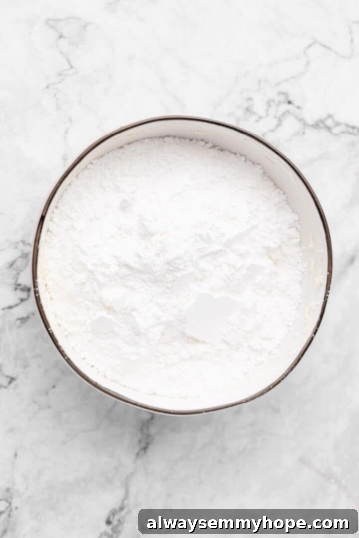 Overhead view of sifted powdered sugar being added to the mixing bowl with creamed vegan butter and cream cheese.