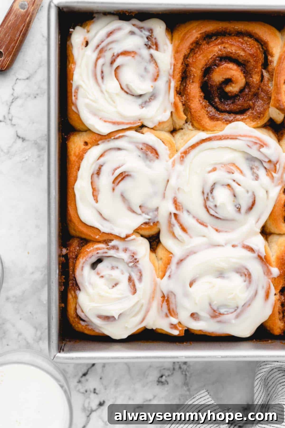 Vegan cinnamon rolls in a baking pan with one unfrosted roll next to a heavily frosted one, showing the before and after.