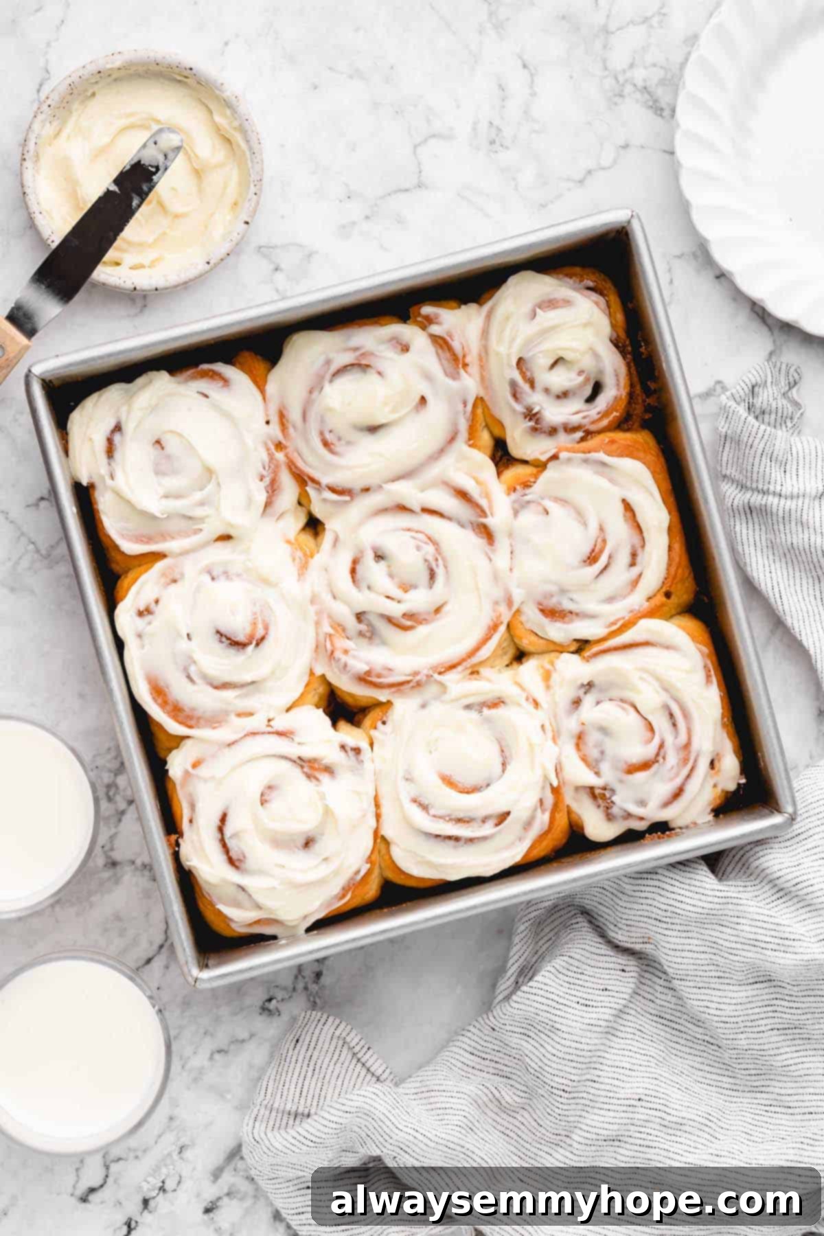Overhead view of freshly baked and frosted vegan cinnamon rolls in a square baking pan, with a bowl of extra frosting nearby.