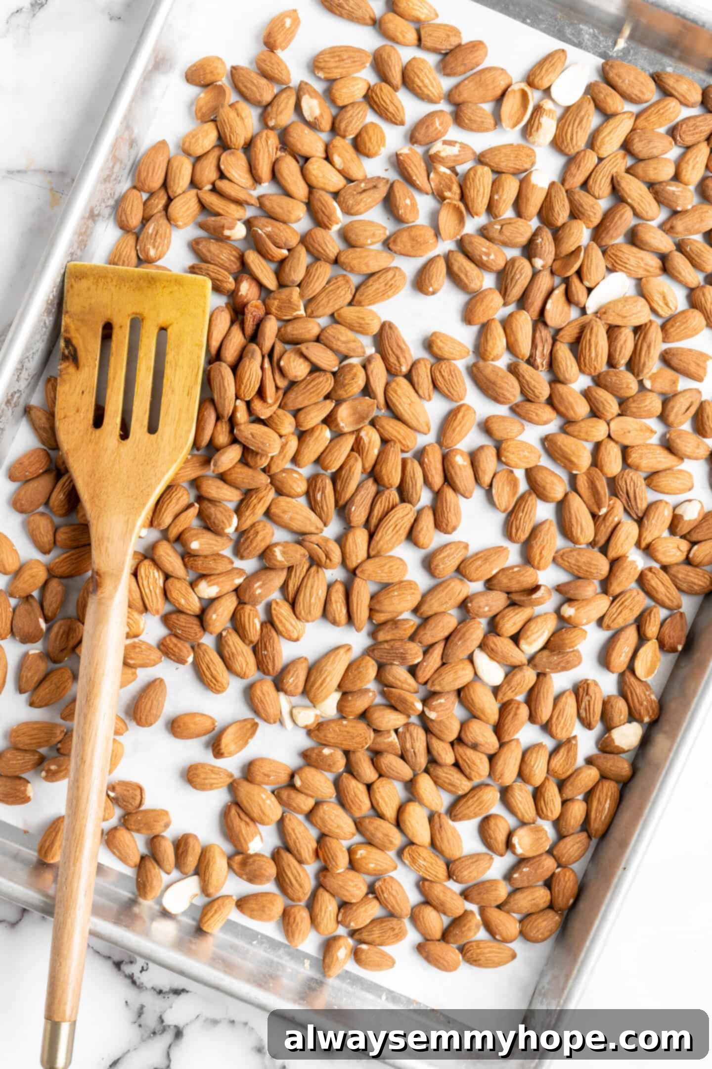 Overhead view of raw almonds spread evenly on a parchment-lined baking sheet, with a wooden spatula nearby