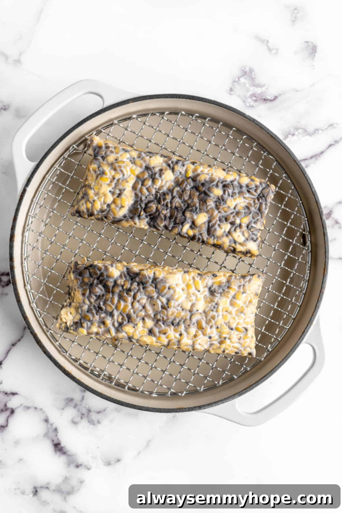 Two blocks of tempeh resting in a steamer basket over simmering water, illustrating the steaming process.