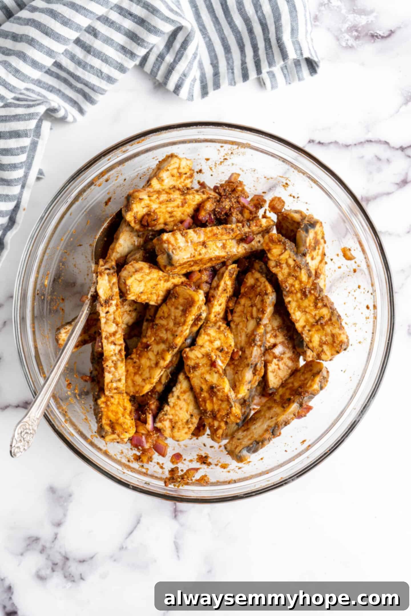 Sliced tempeh strips generously tossed and coated in the savory marinade within a mixing bowl.