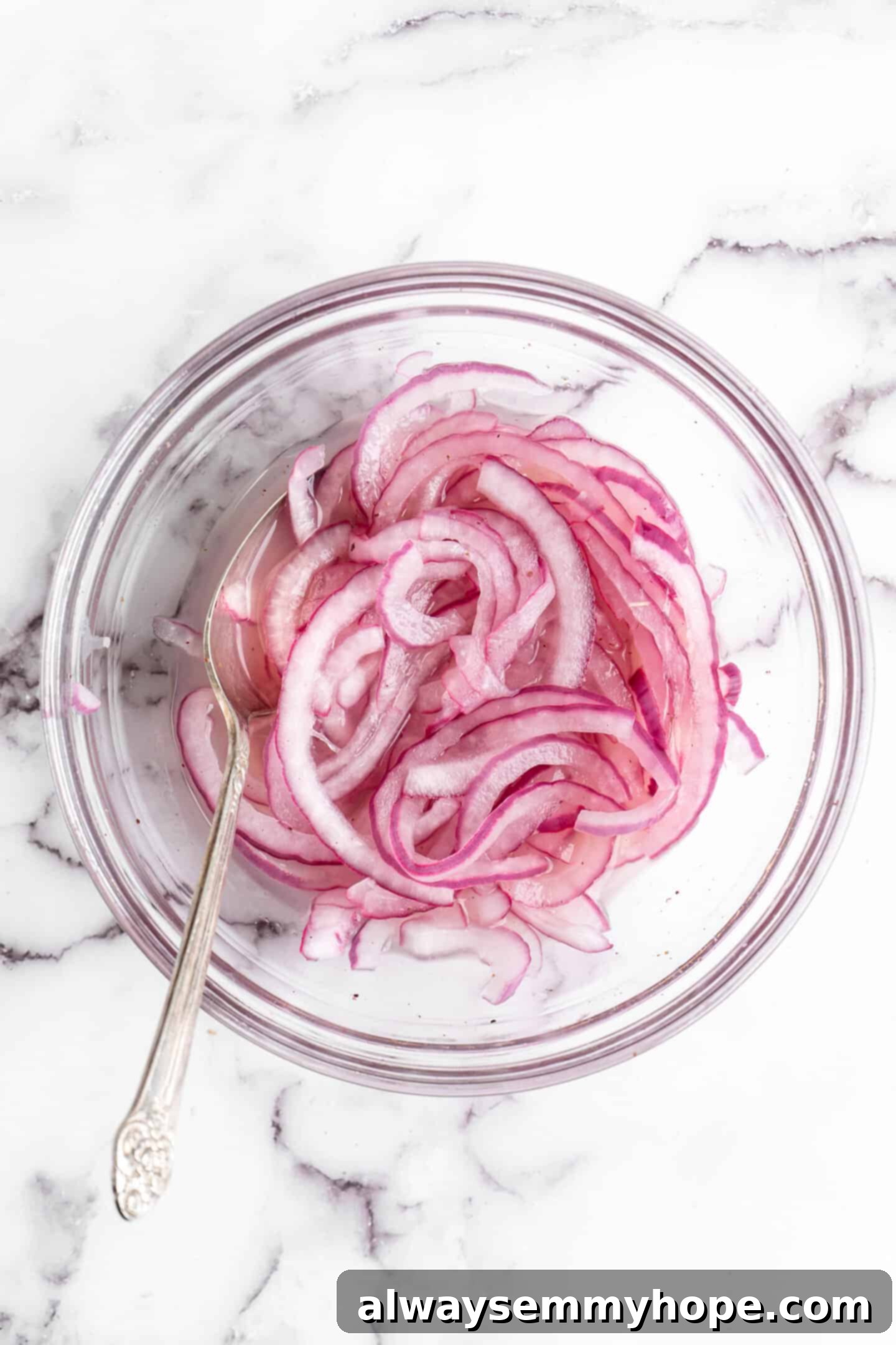 A glass bowl filled with vibrant quick-pickled red onions, accompanied by a spoon for serving.