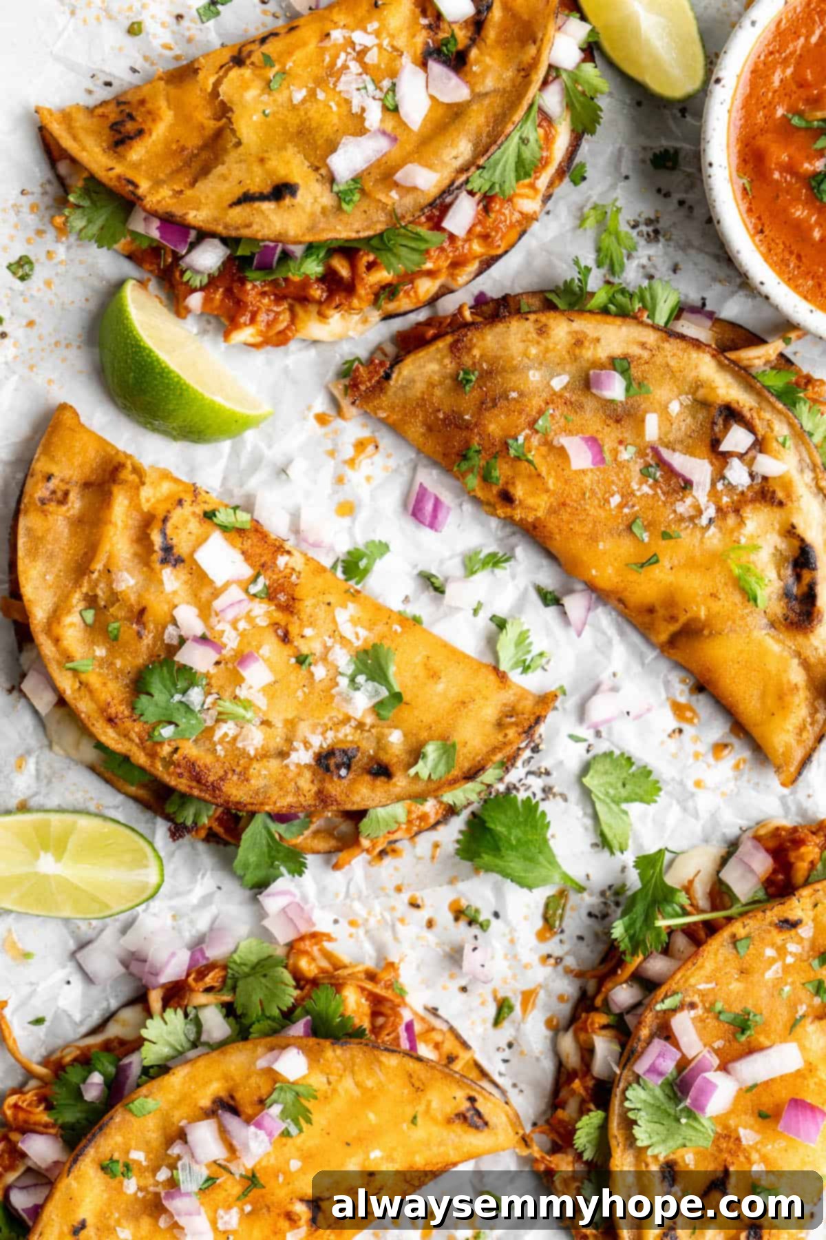 Vegan birria tacos use king oyster mushrooms to mimic the texture/flavour of the original. They’re smoky, crispy, and perfect for dipping! Overhead view of vegan birria tacos with garnishes.