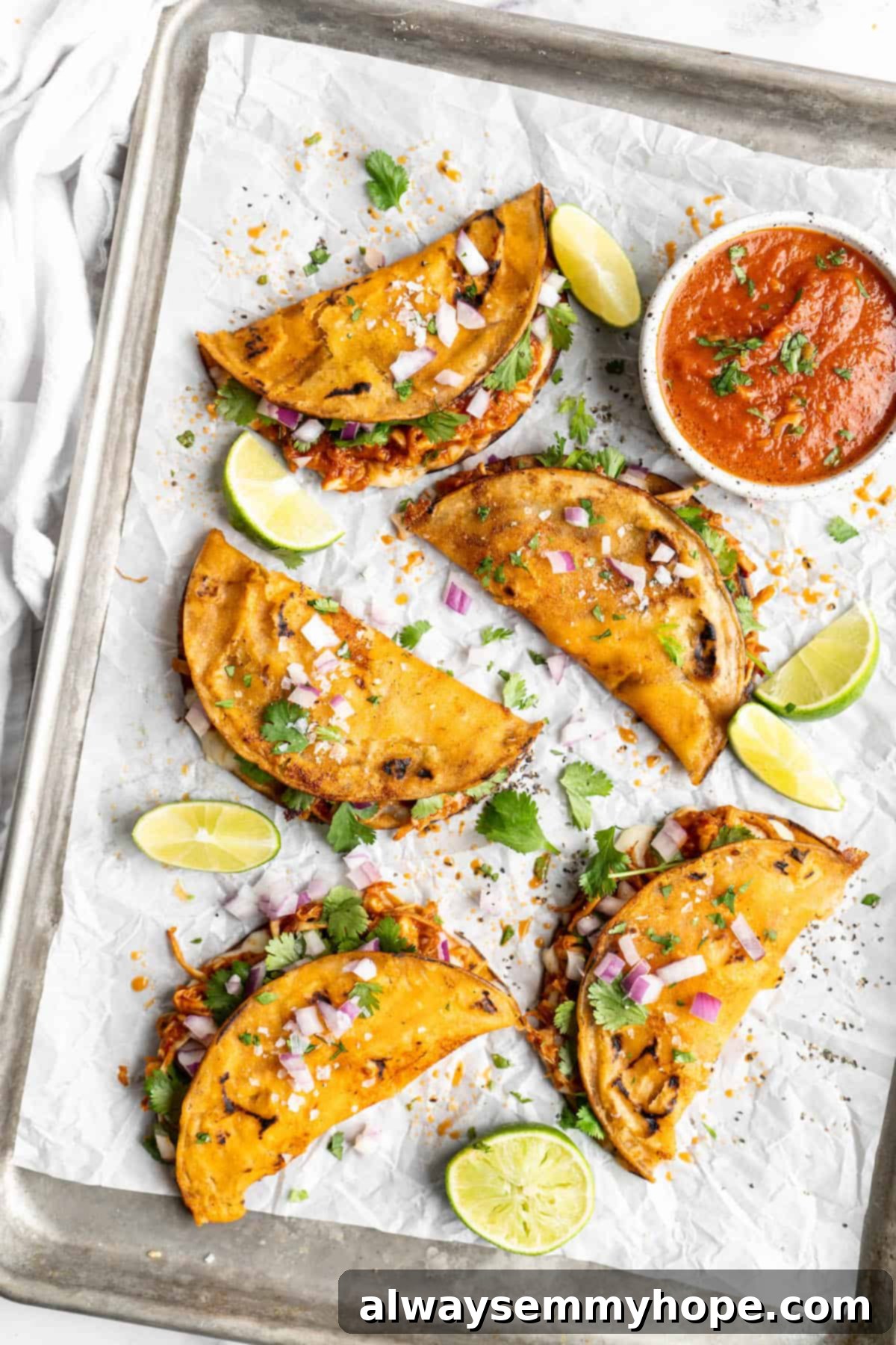 Vegan birria tacos use king oyster mushrooms to mimic the texture/flavour of the original. They’re smoky, crispy, and perfect for dipping! Overhead view of vegan birria tacos on a sheet pan with lime wedges and a bowl of sauce.
