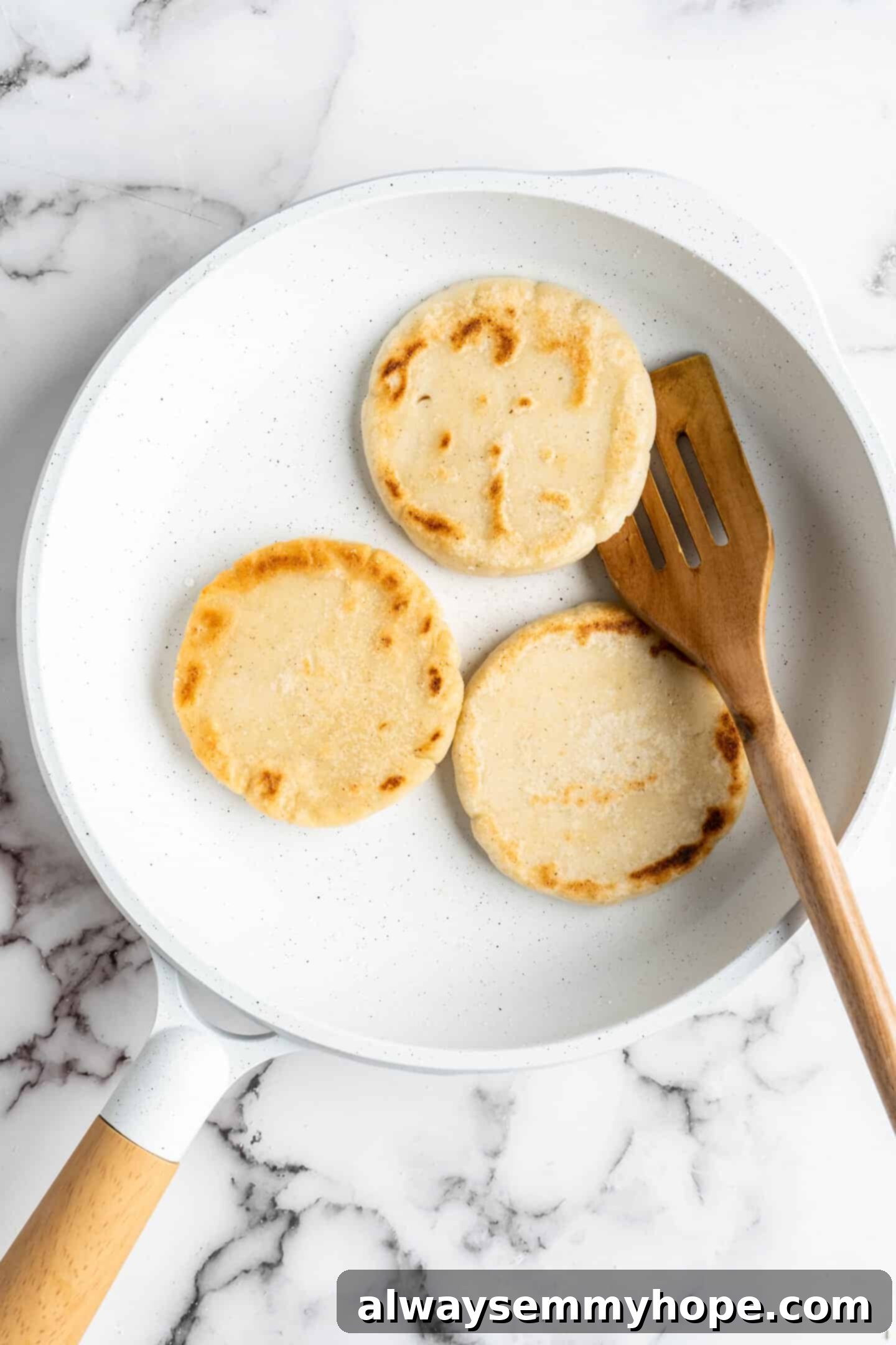 This step-by-step tutorial will show you how to make perfect vegan arepas with just a few ingredients. They're easier to make than you think! Overhead view of three vegan arepas sizzling in a skillet, with a wooden spatula poised to flip one, showing the golden-brown crust.