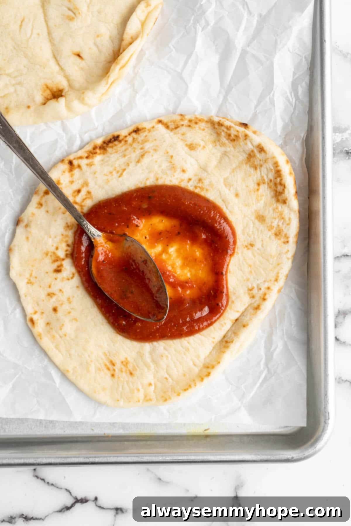 Overhead view of a spoon spreading rich tomato pizza sauce evenly onto a vegan naan base.