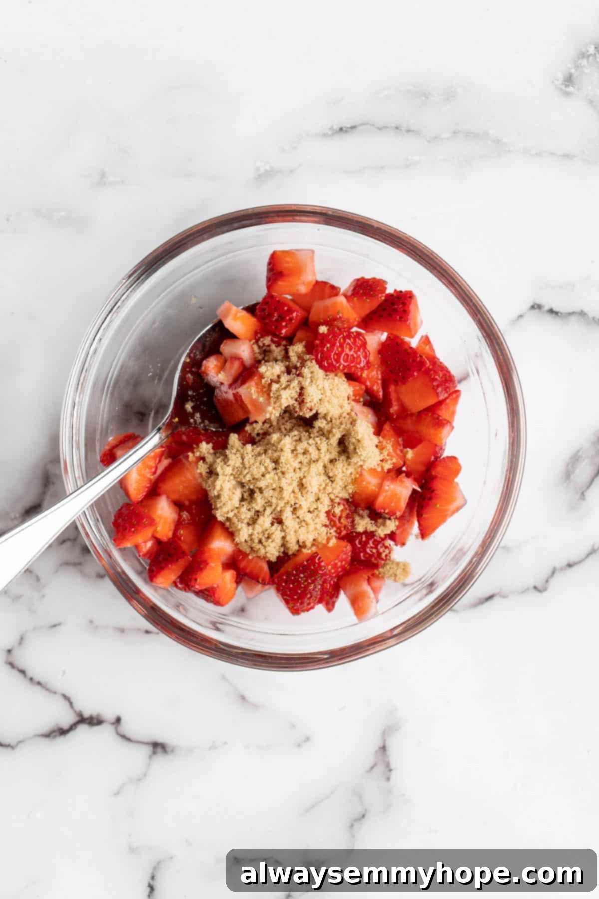 How to make vegan Korean strawberry milk! This easy recipe is made with 4 simple ingredients and each glass is full of fresh strawberries. Overhead view of diced fresh strawberries being mixed with brown sugar in a clear glass bowl, beginning the maceration process.