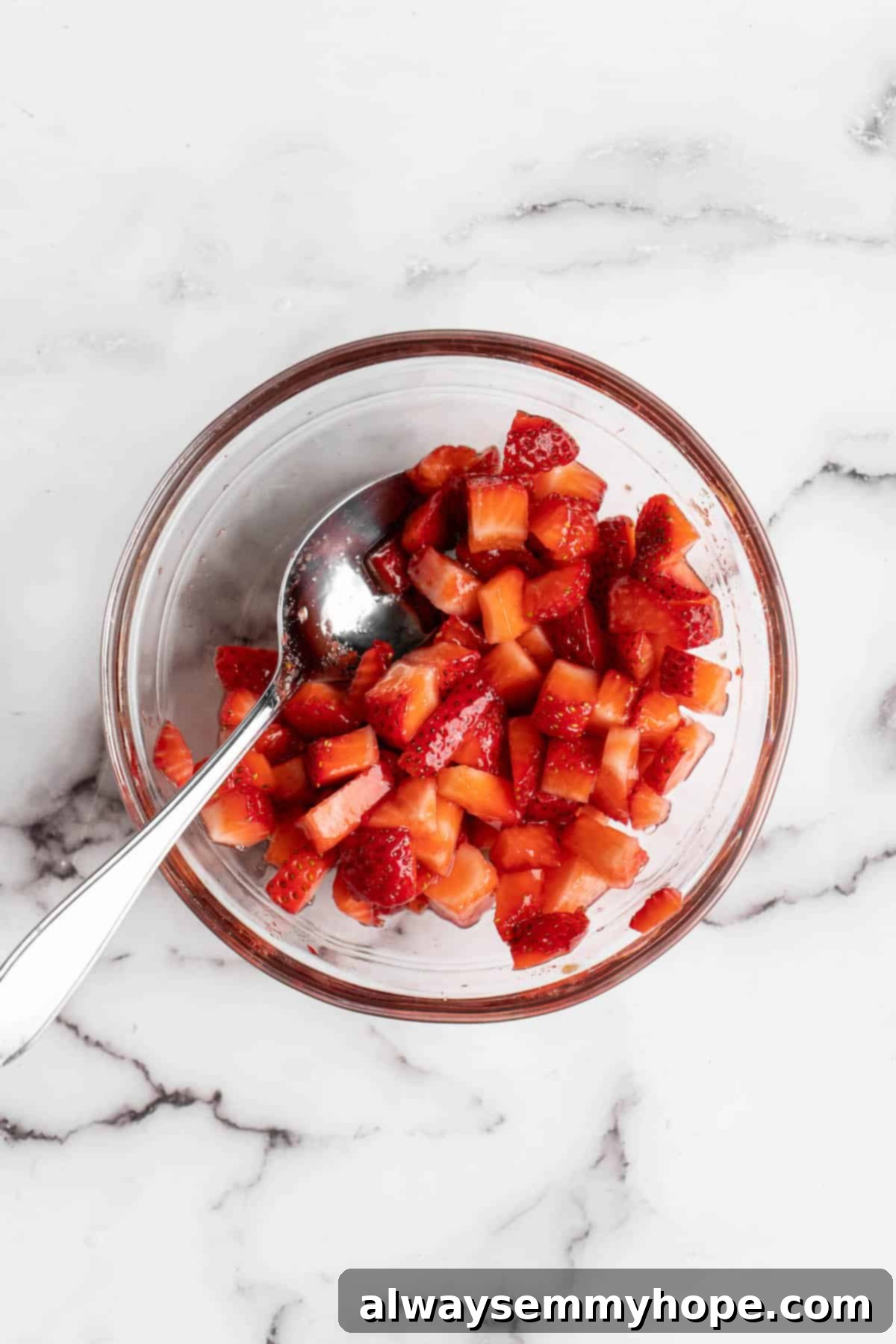 How to make vegan Korean strawberry milk! This easy recipe is made with 4 simple ingredients and each glass is full of fresh strawberries. Overhead view of macerated strawberries resting in a clear glass bowl, having released their juices after being mixed with sugar.