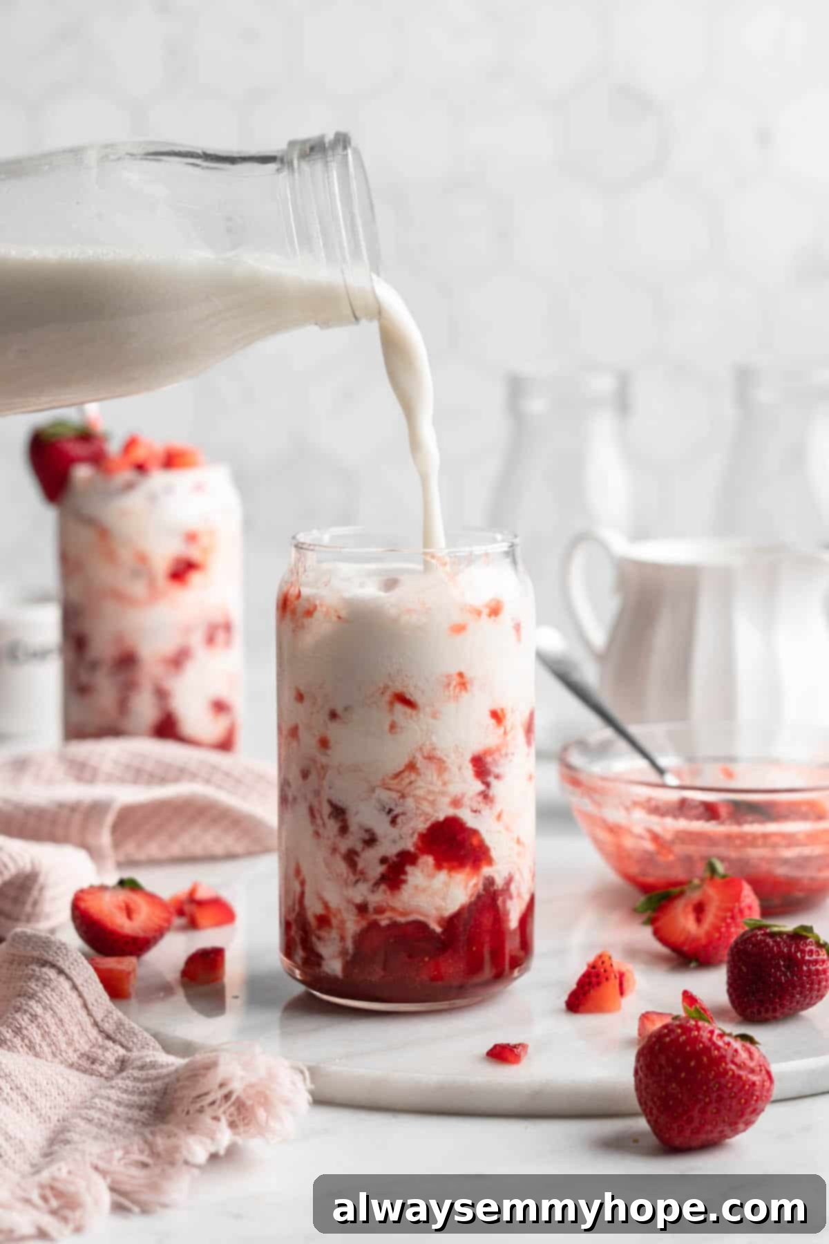 How to make vegan Korean strawberry milk! This easy recipe is made with 4 simple ingredients and each glass is full of fresh strawberries. A hand pouring creamy plant-based milk into a glass already containing strawberry syrup and macerated strawberries, creating distinct layers.