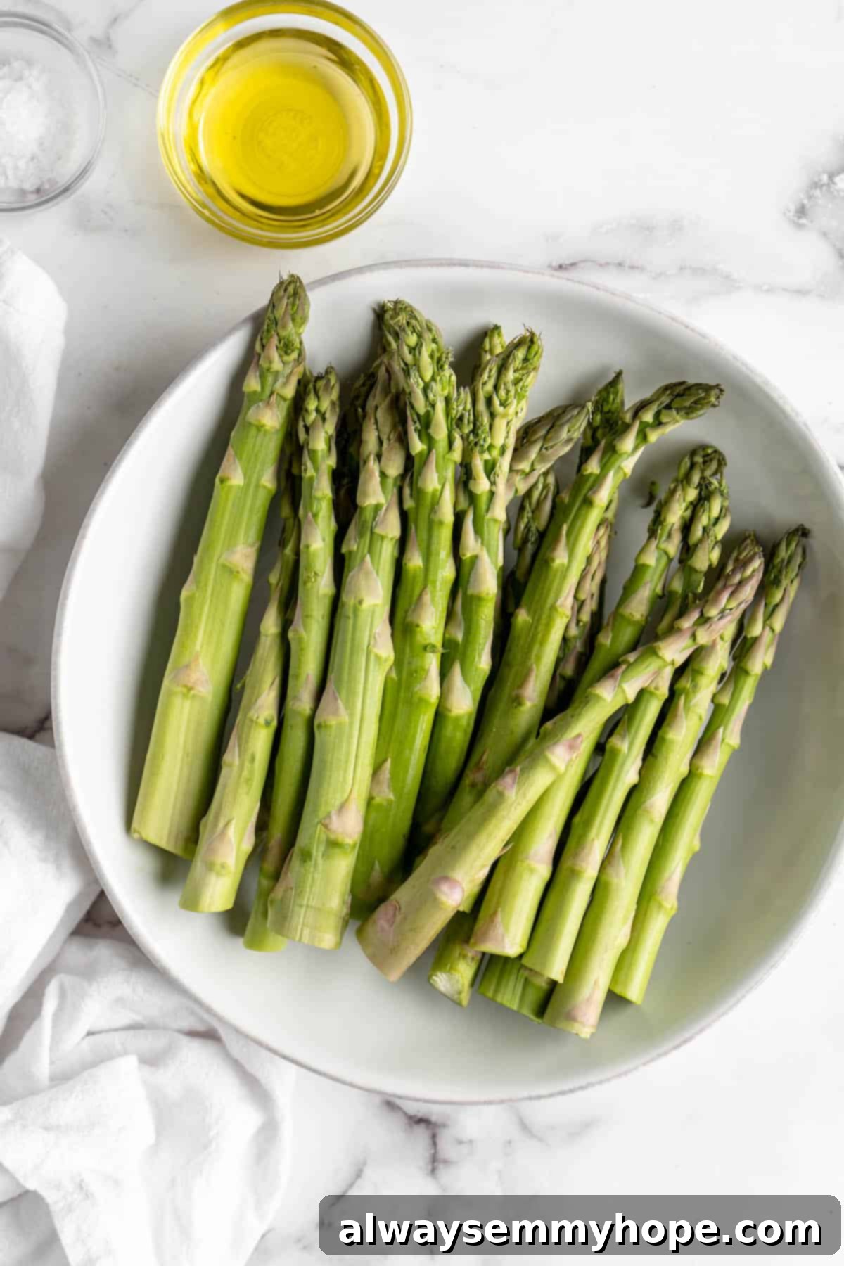 This easy lemon pepper air fryer asparagus is the perfect side dish for busy weeknights. It's done in just 10 minutes from start to finish! Overhead view of fresh, trimmed asparagus in a mixing bowl with a small bowl of olive oil on the side.