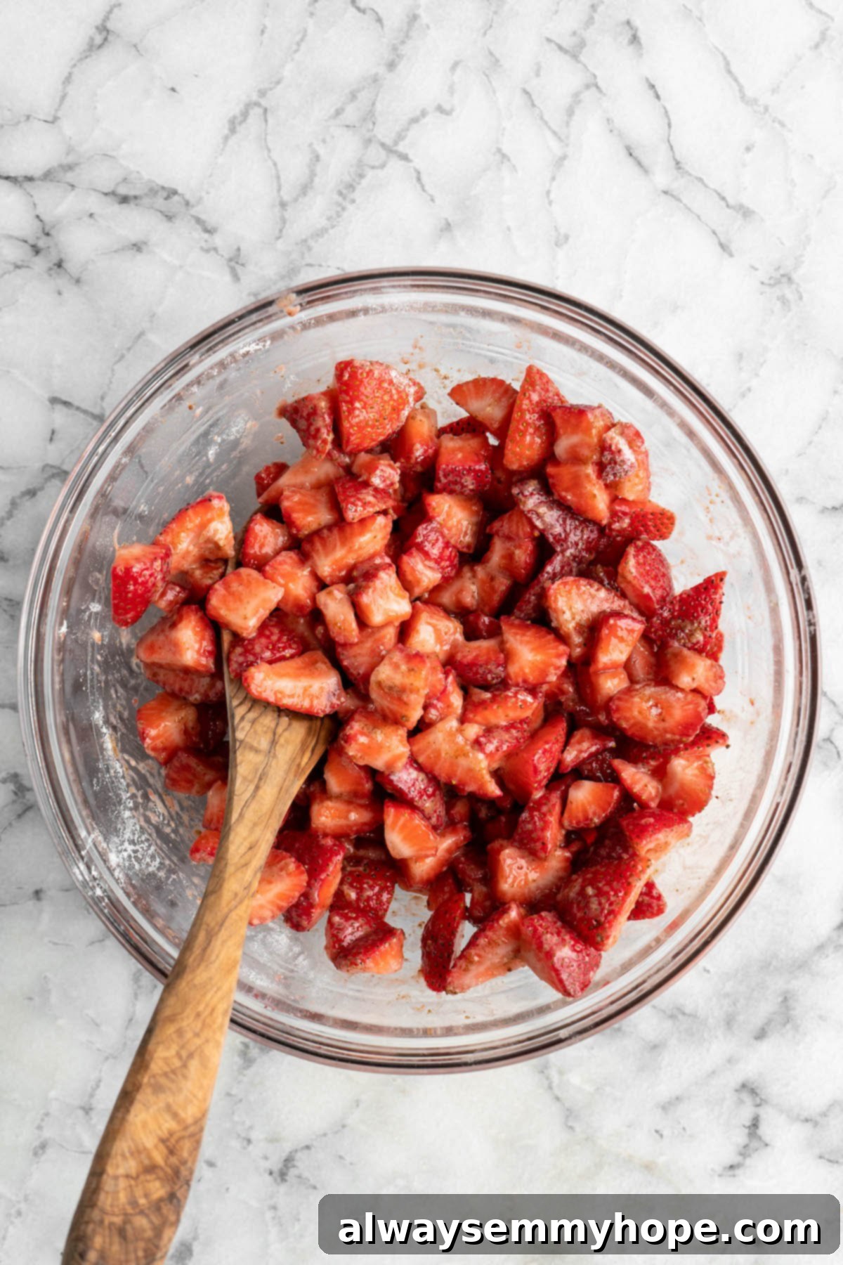 This vegan strawberry crumble is easy as can be! You only need 5 ingredients and a few minutes of assembly for the perfect summer dessert. Overhead view of macerated strawberries in glass bowl with wooden spoon