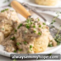 A plate with two biscuits and gravy on it, garnished with chives, with a fork and knife on the plate and biscuits in the background