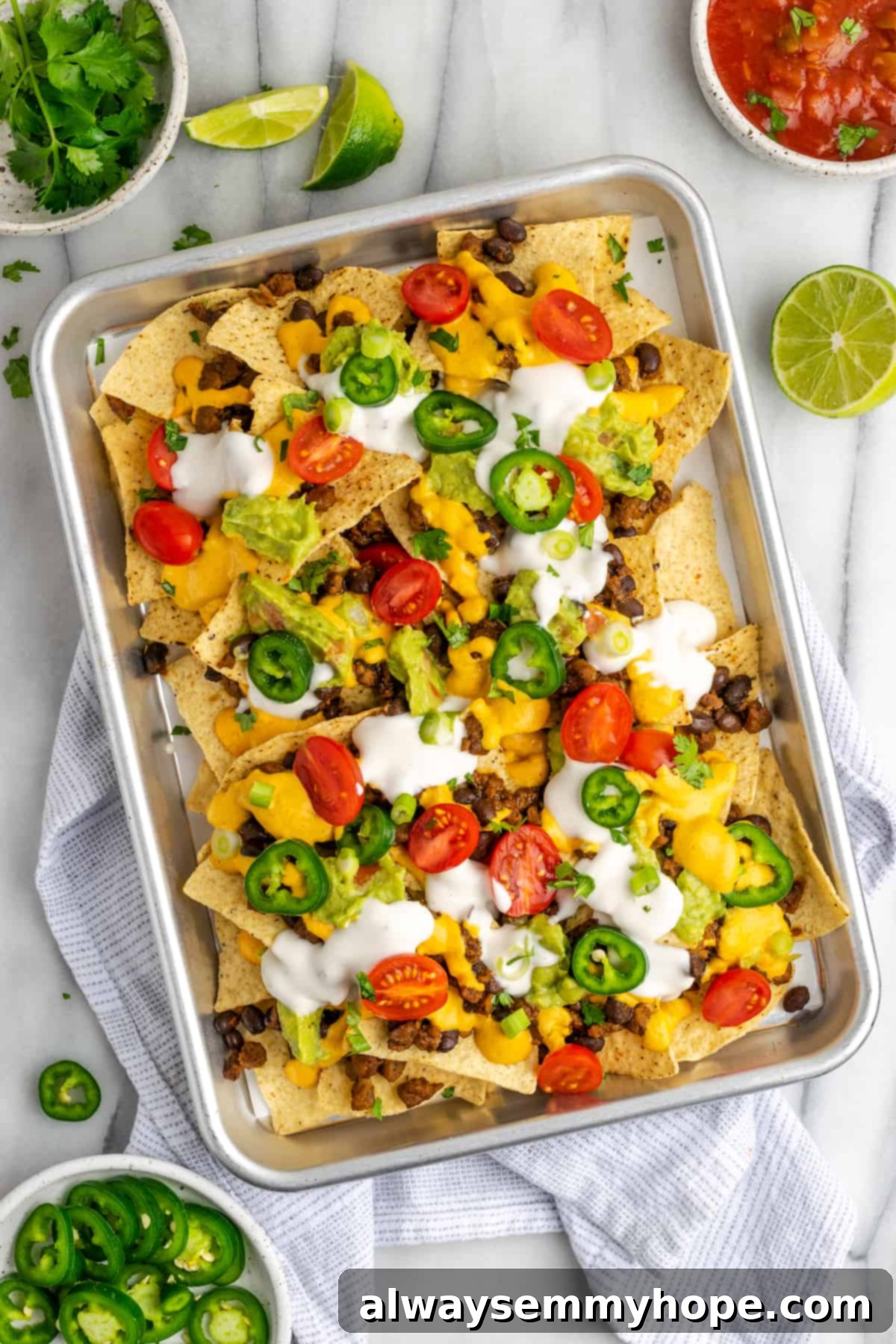 These vegan loaded nachos are rich and filling, with a spicy vegan meat crumble, black beans, a gooey vegan cheese sauce, and toppings. Overhead view of a tray of loaded nachos, covered in vegan sour cream, jalapeño slices, tomatoes, and cilantro, next to a half a lime, a bowl of cilantro, a bowl of jalapeños, and a bowl of salsa.