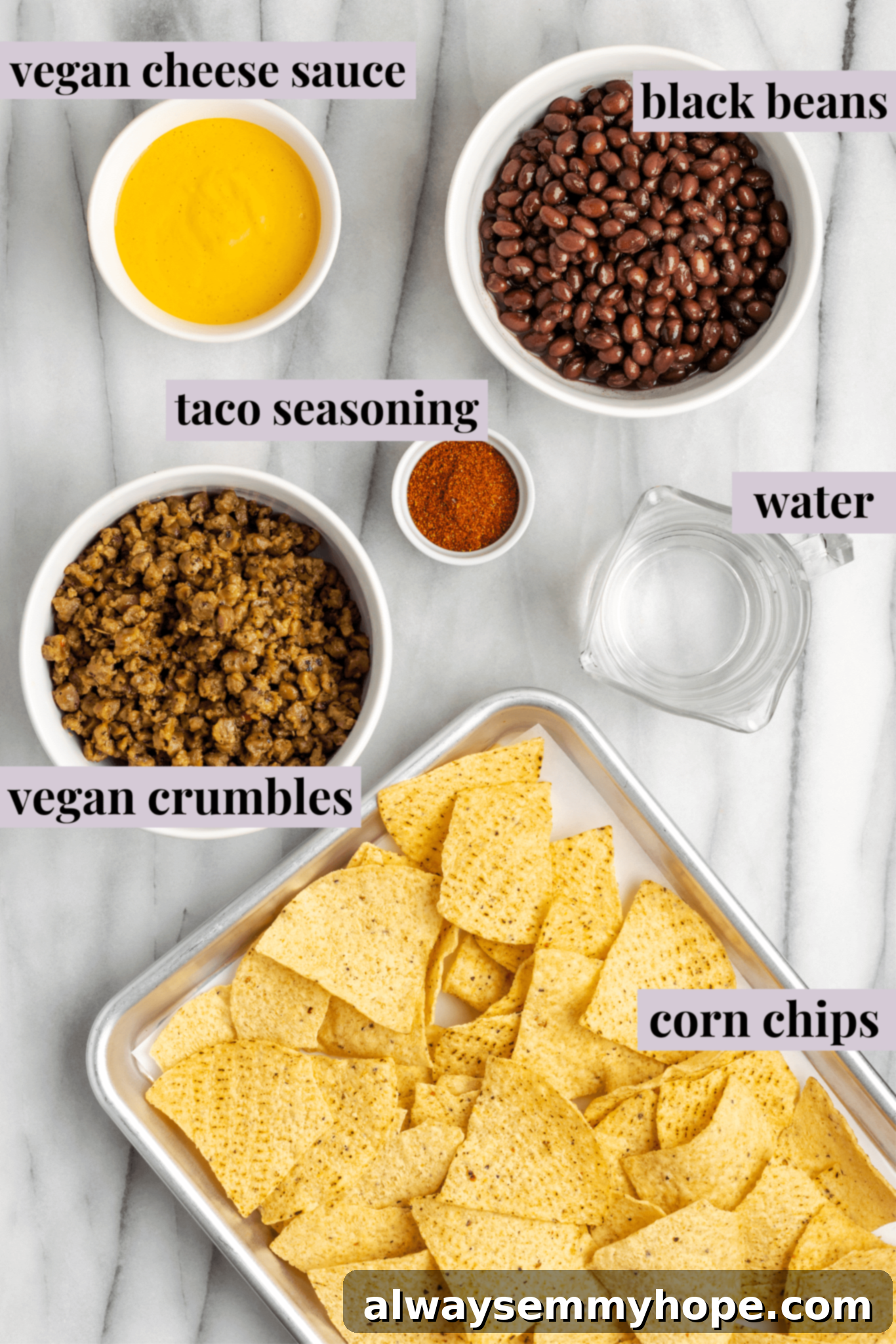 These vegan loaded nachos are rich and filling, with a spicy vegan meat crumble, black beans, a gooey vegan cheese sauce, and toppings. Overhead view of a tray of tortilla chips, a bowl of vegan meat crumbles, a bowl of black beans, a bowl of vegan cheese sauce, a bowl of taco seasoning, and a glass of water.