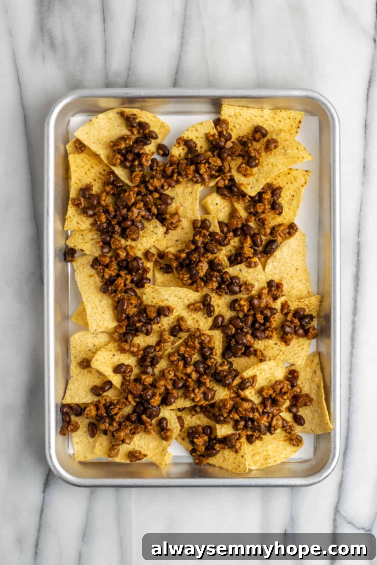 These vegan loaded nachos are rich and filling, with a spicy vegan meat crumble, black beans, a gooey vegan cheese sauce, and toppings. A baking tray with a layer of tortilla chips, topped with a mixture of black beans and vegan meat crumbles.