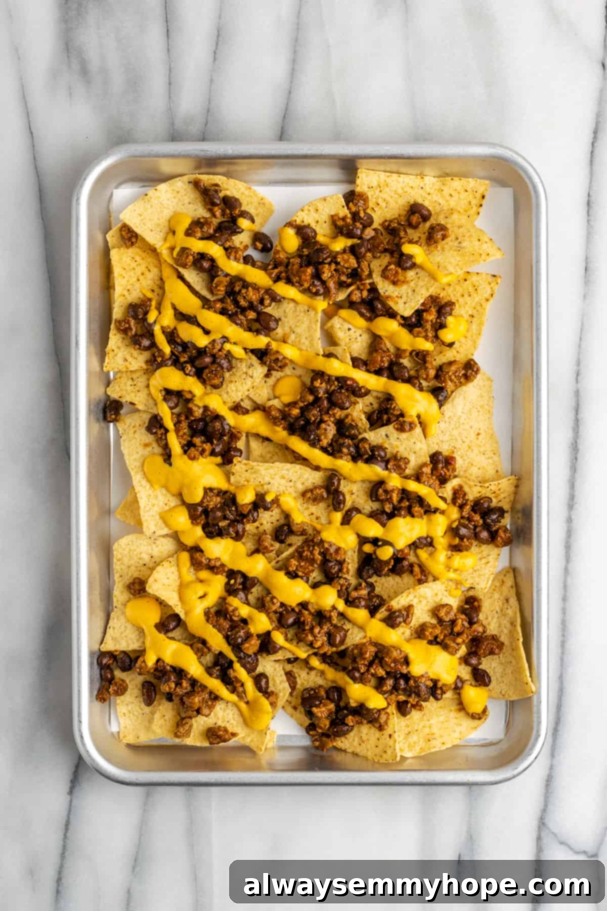 These vegan loaded nachos are rich and filling, with a spicy vegan meat crumble, black beans, a gooey vegan cheese sauce, and toppings. A baking sheet with a layer of chips, topped with vegan meat crumbles and beans, with vegan cheese sauce poured on top.