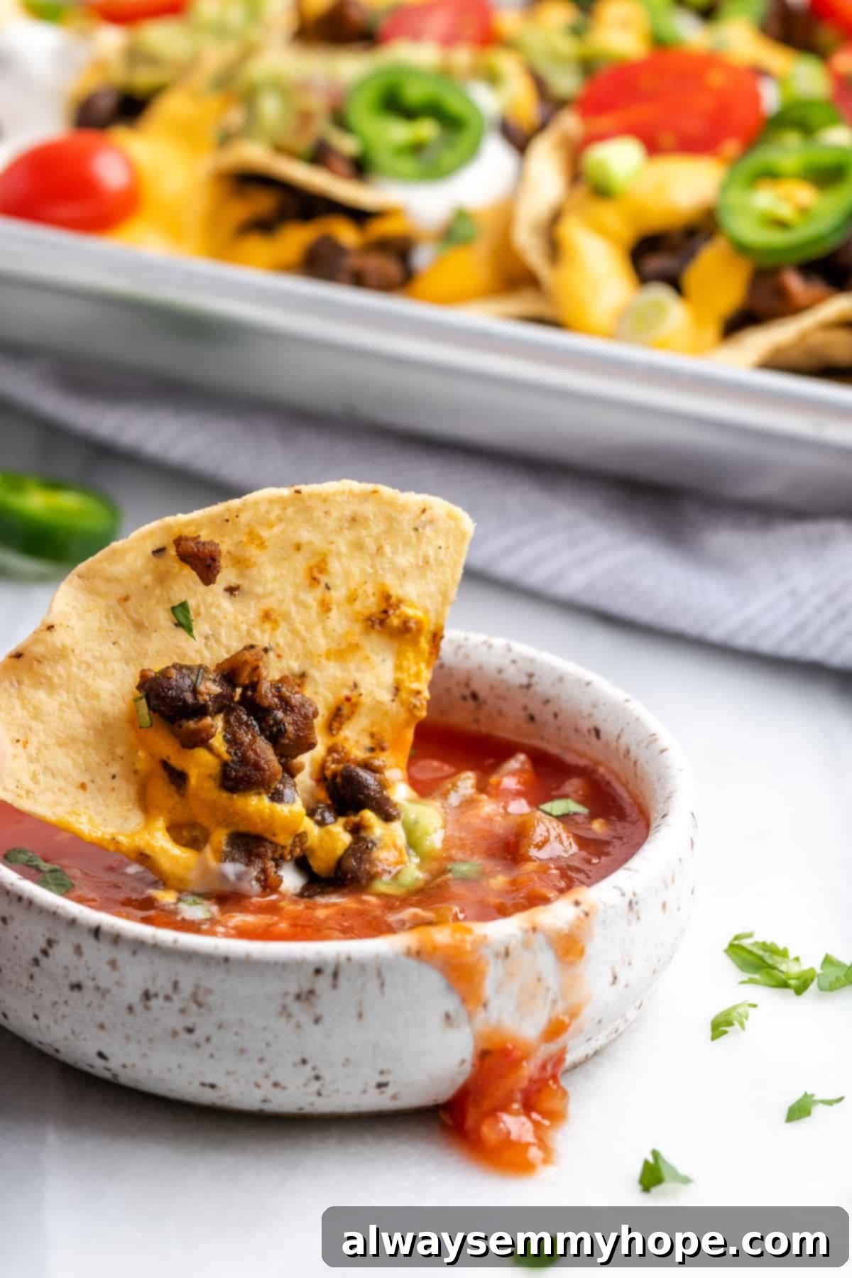 These vegan loaded nachos are rich and filling, with a spicy vegan meat crumble, black beans, a gooey vegan cheese sauce, and toppings. A chip with beans and cheese on it dipping into a bowl of salsa, with a tray of nachos in the background.