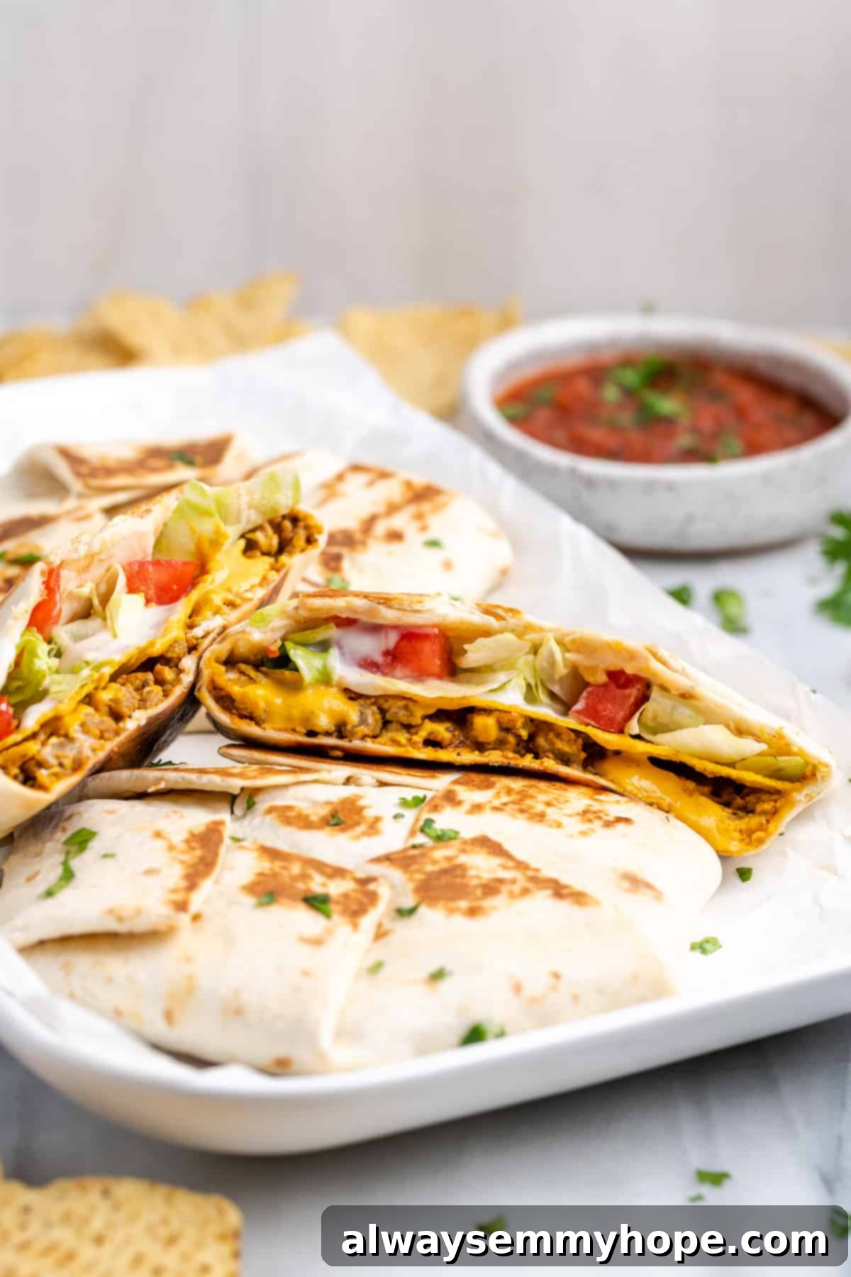 A rectangular plate with a vegan crunchwrap supreme cut in half on top of other crunchwraps, with a bowl of salsa in the background. The crunchwrap reveals layers of vegan meat, cheese, tostada, and fresh vegetables.