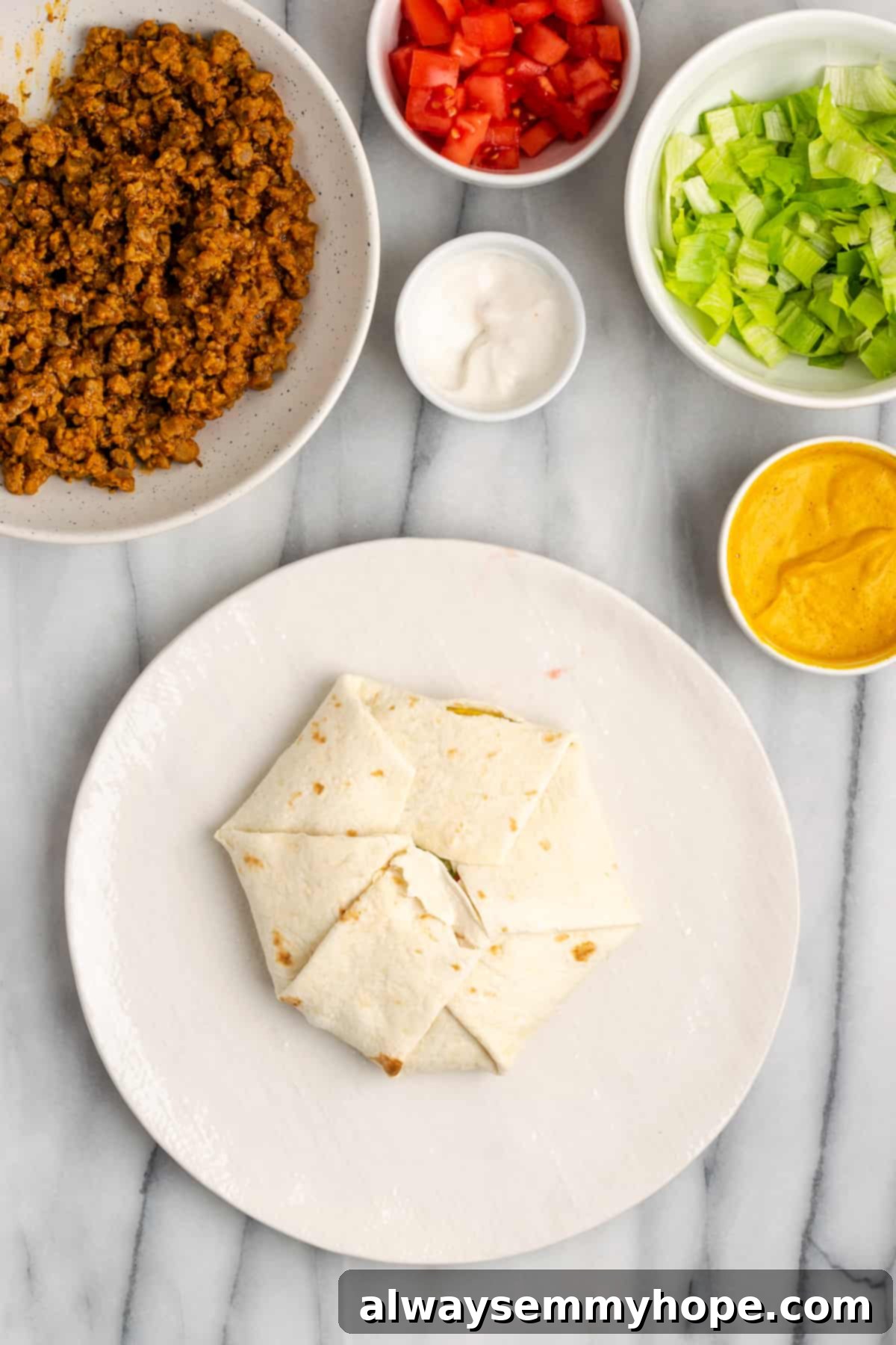 A plate with an untoasted, neatly folded vegan crunchwrap supreme, ready for the skillet. Bowls of remaining ingredients are in the background.