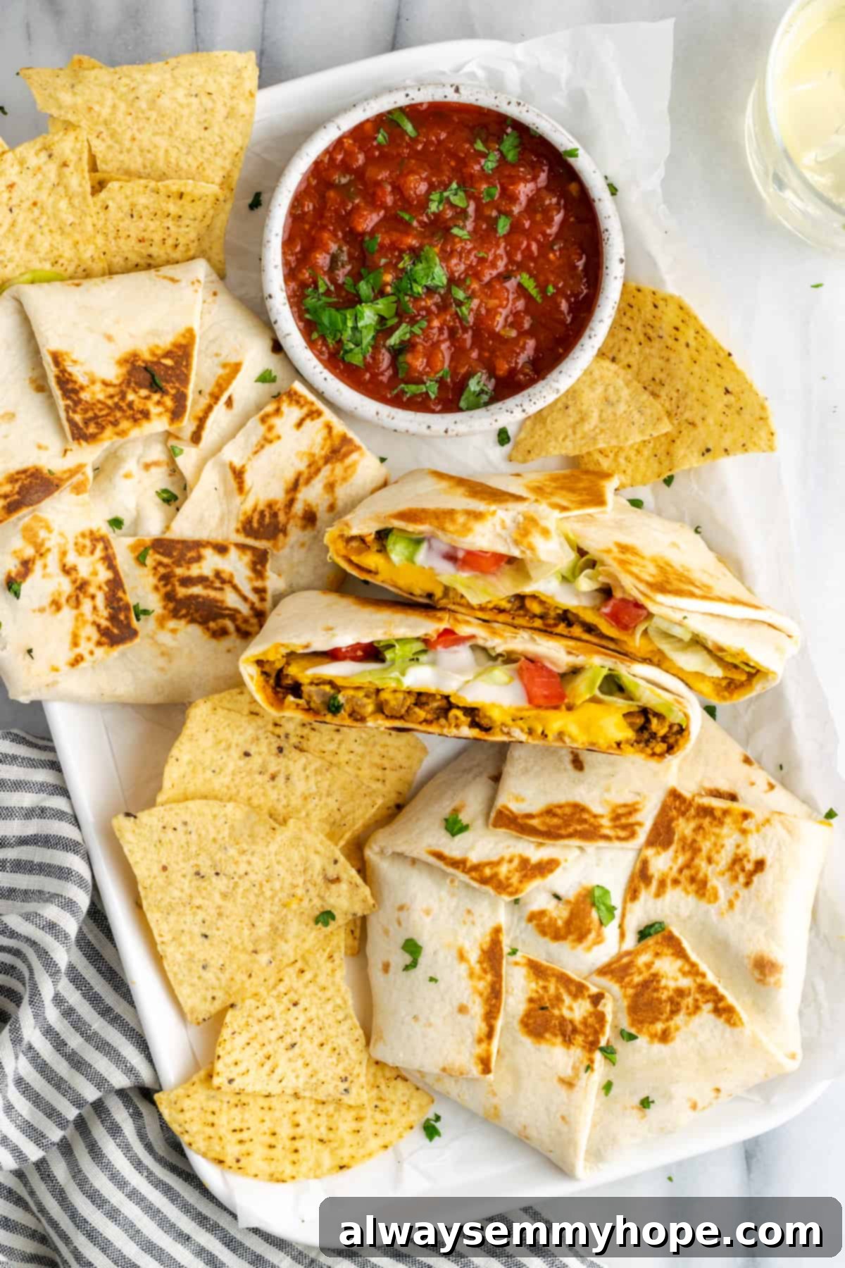 A plate with two whole vegan crunchwrap supremes, one crunchwrap supreme cut in half to show the layers, some tortilla chips, and a small bowl of fresh salsa for dipping. The meal looks inviting and complete.