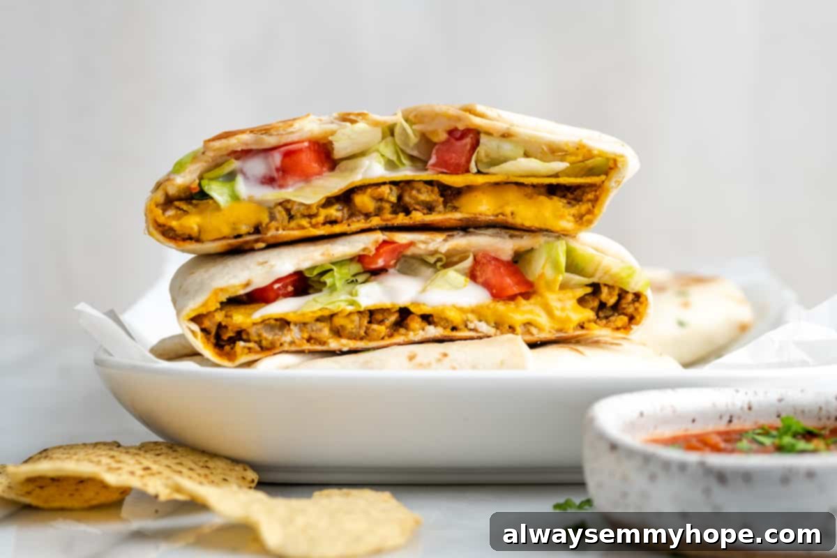 A vegan crunchwrap supreme expertly cut in half, with the two sides stacked to showcase the perfect layers of vegan meat, cheese, tostada, sour cream, lettuce, and tomato. It rests on a whole crunchwrap supreme, surrounded by crunchy tortilla chips.
