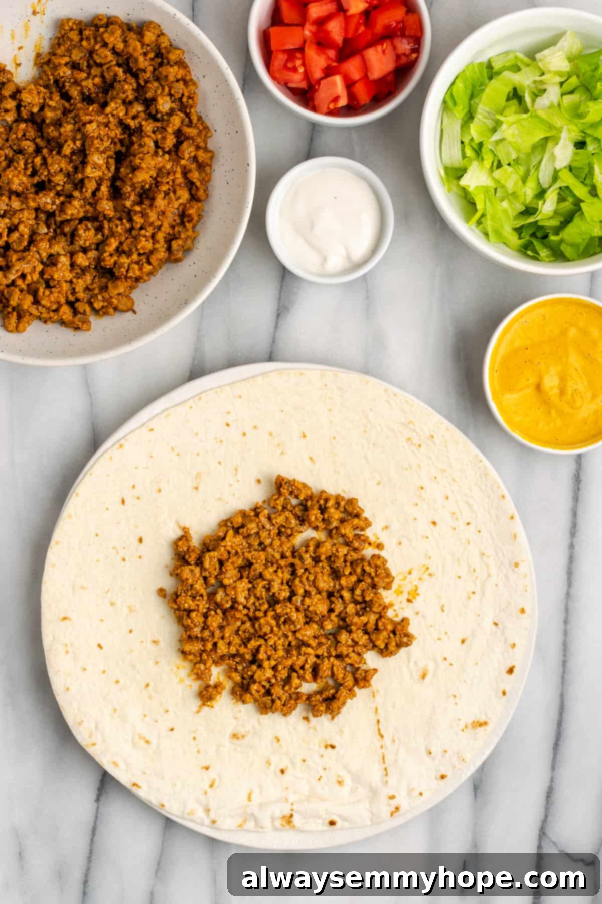 A flour tortilla carefully topped with a generous layer of seasoned vegan meat crumbles, forming a circular base for the crunchwrap. Bowls of other fillings are visible in the background.