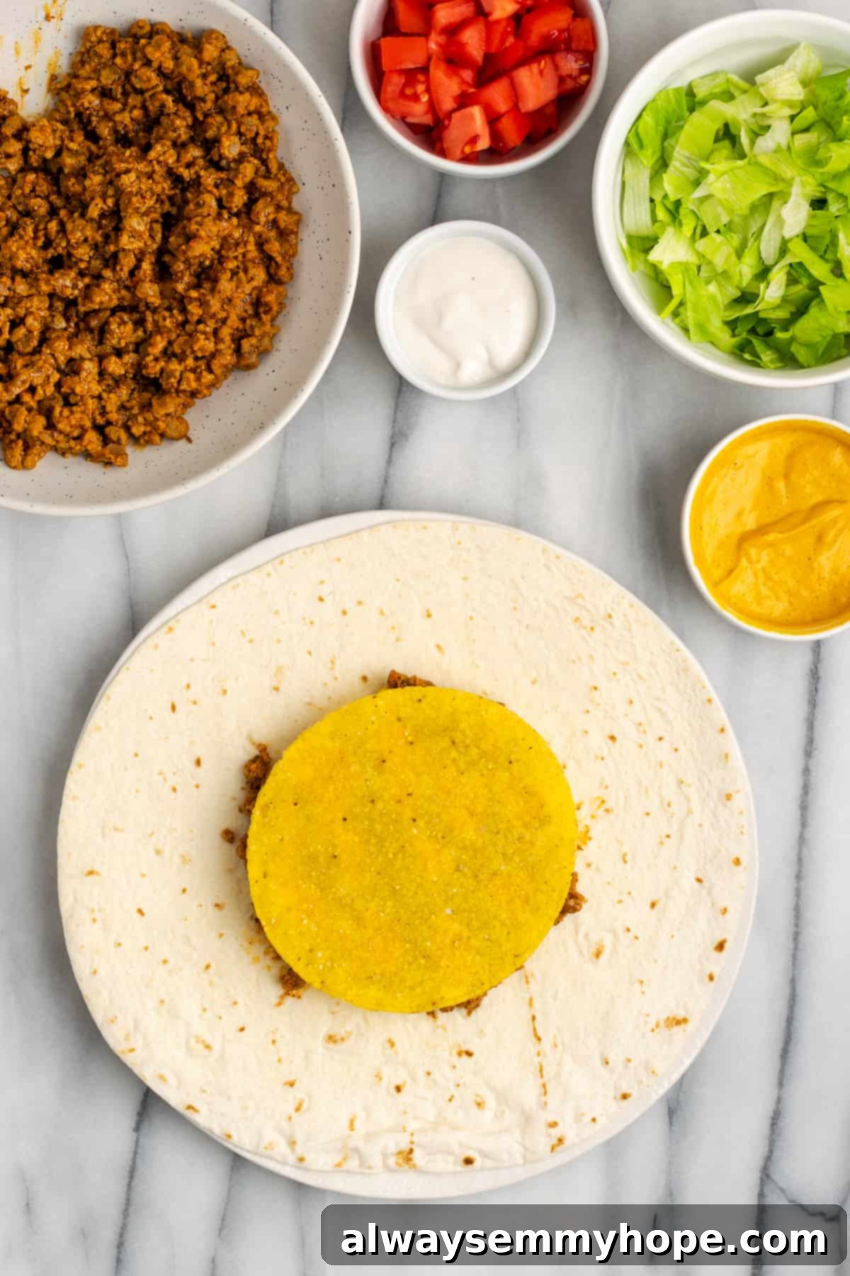 A flour tortilla layered with vegan meat, cheese sauce, and a crisp tostada shell, forming the structural core of the crunchwrap. Surrounding bowls hold fresh toppings.