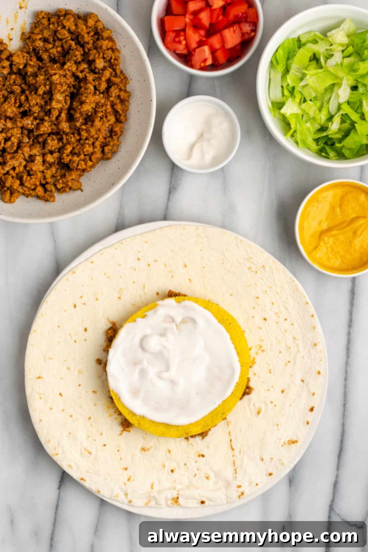 A flour tortilla assembly now includes vegan meat, cheese sauce, a tostada, and a smooth layer of vegan sour cream. The colors are vibrant and inviting.