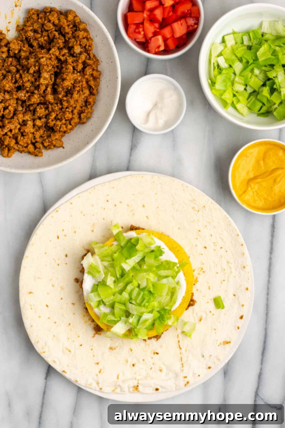 A partially assembled crunchwrap with vegan meat, cheese, tostada, sour cream, and a layer of crisp shredded lettuce. All the fresh ingredients are ready for the final touches.