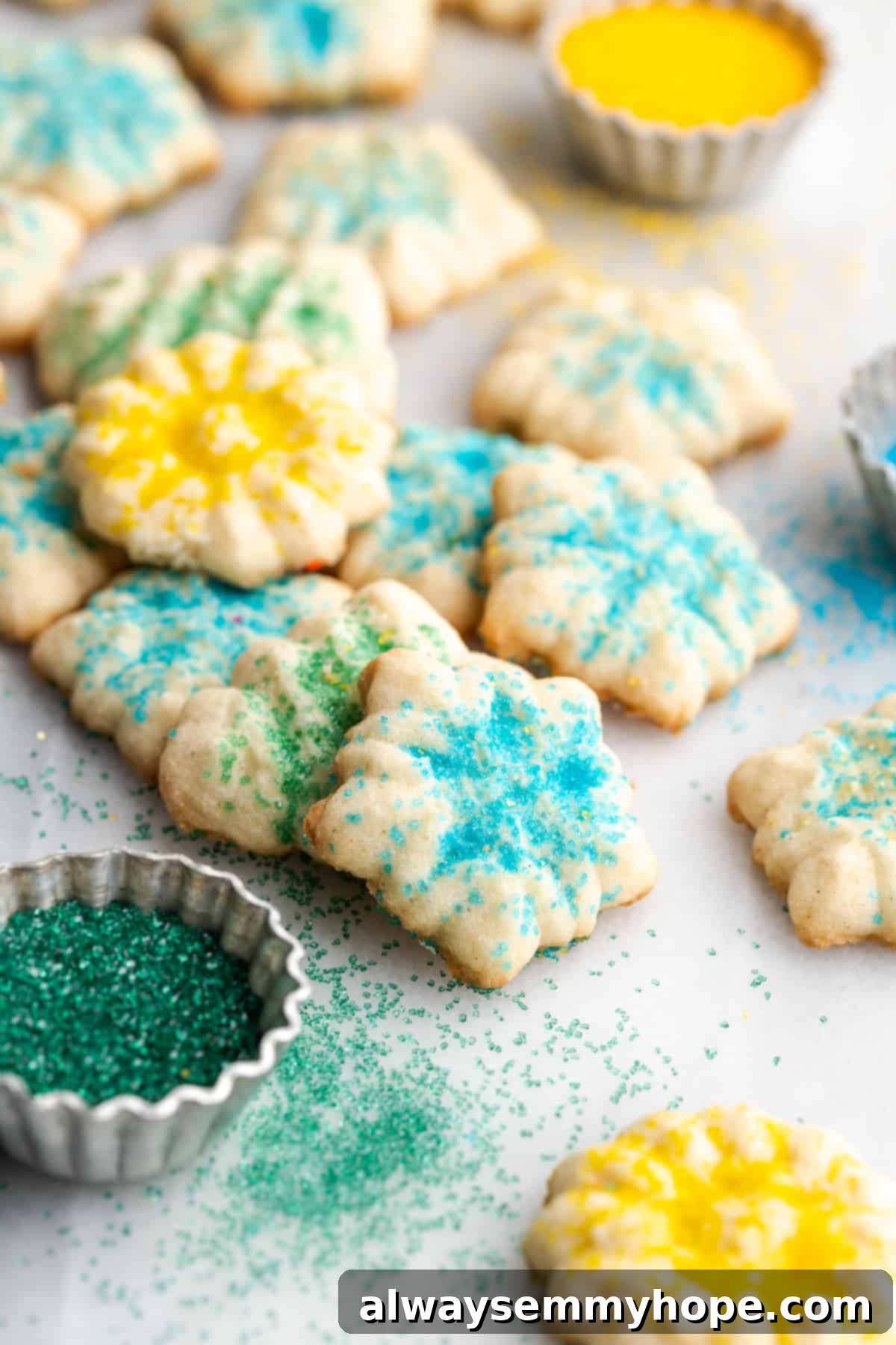 This buttery vegan spritz cookies recipe is fun to make and eat, and the cookies are moist, tender, and full of almond and vanilla flavor. A pile of spritz cookies with various sprinkles on them, next to a bowl of green sprinkles and a bowl of yellow sprinkles, showcasing their festive appeal