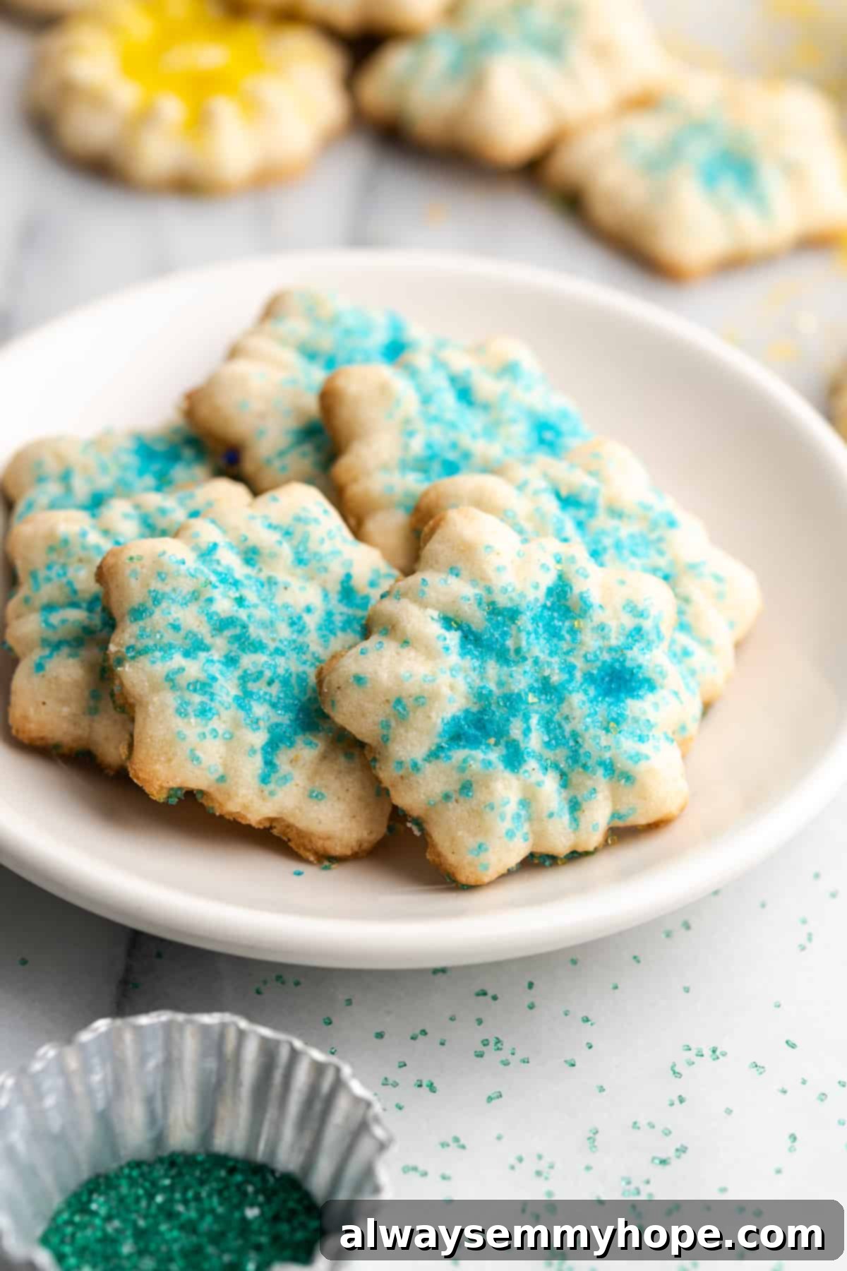 This buttery vegan spritz cookies recipe is fun to make and eat, and the cookies are moist, tender, and full of almond and vanilla flavor. A plate filled with spritz cookies covered in blue sprinkles, with cookies in the background with different sprinkles, ready to be enjoyed