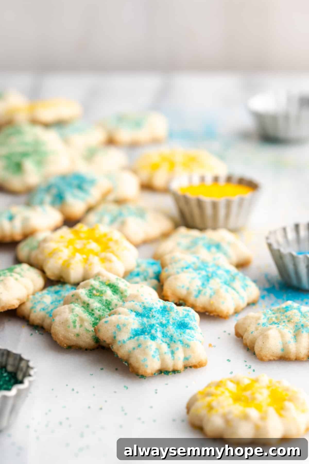 This buttery vegan spritz cookies recipe is fun to make and eat, and the cookies are moist, tender, and full of almond and vanilla flavor. Lots of spritz cookies covered in sprinkles, next to a bowl of yellow sprinkles, emphasizing abundance and variety