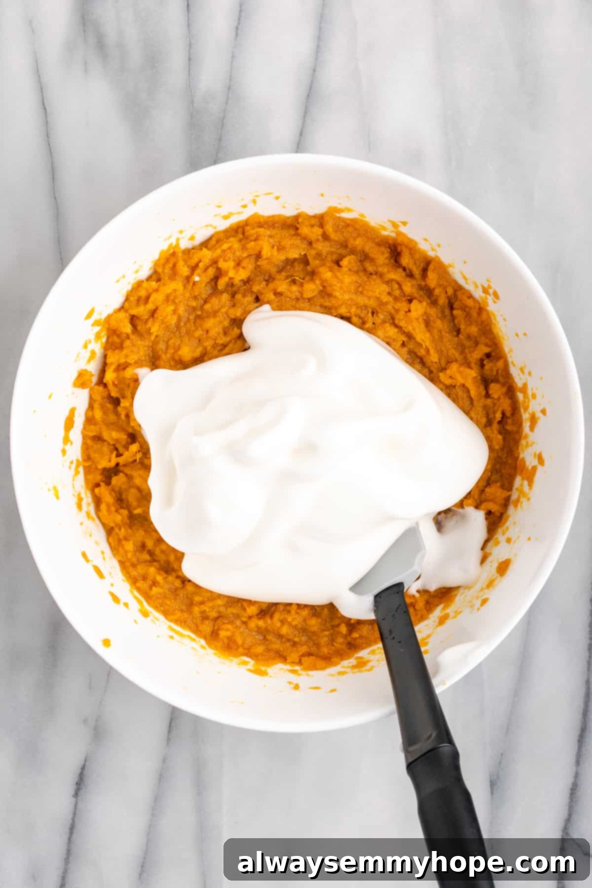 This rich and creamy vegan sweet potato soufflé recipe is a cozy Thanksgiving side. It's entirely vegan, and can easily be made gluten-free. A mixing bowl containing the sweet potato soufflé batter, with a dollop of whipped aquafaba resting on top, and a spatula ready to gently fold it in.