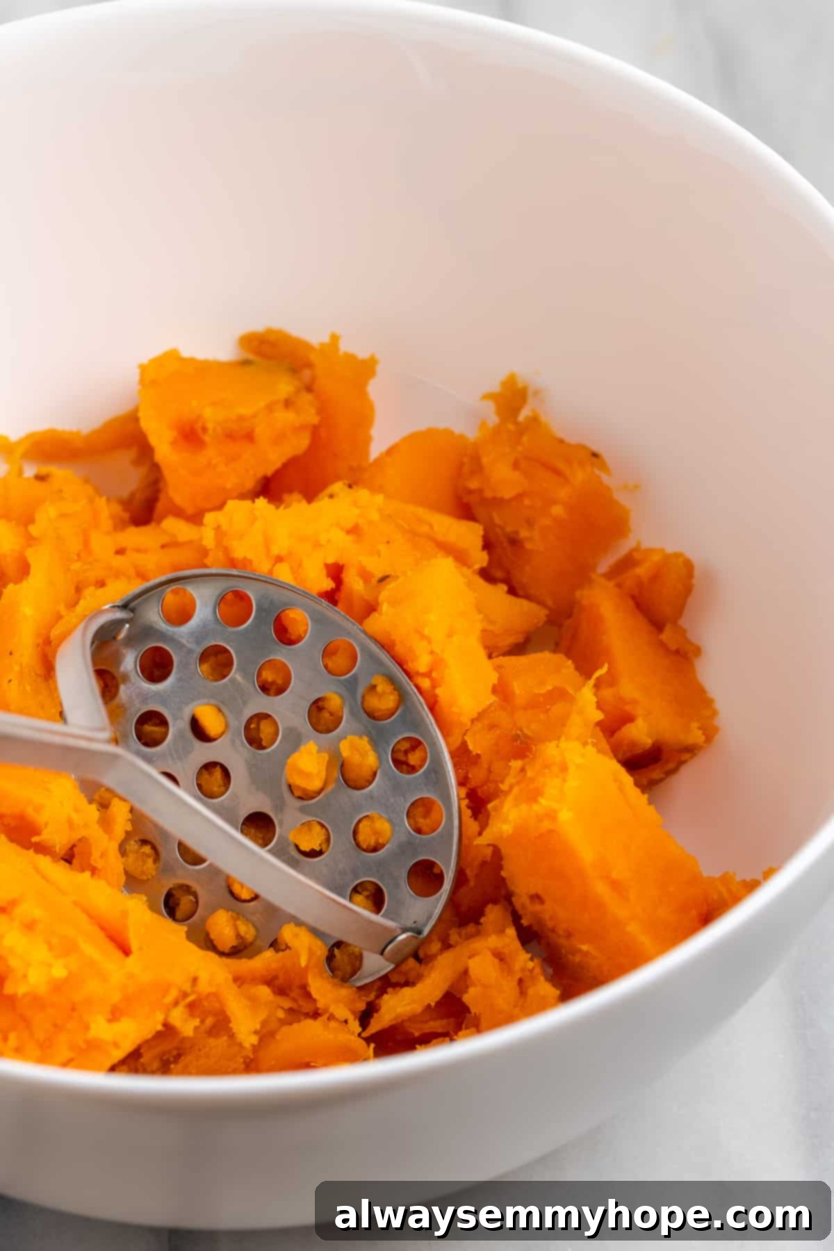 This rich and creamy vegan sweet potato soufflé recipe is a cozy Thanksgiving side. It's entirely vegan, and can easily be made gluten-free. Close-up view of chunks of cooked sweet potato in a mixing bowl, with a potato masher beginning to break them down.