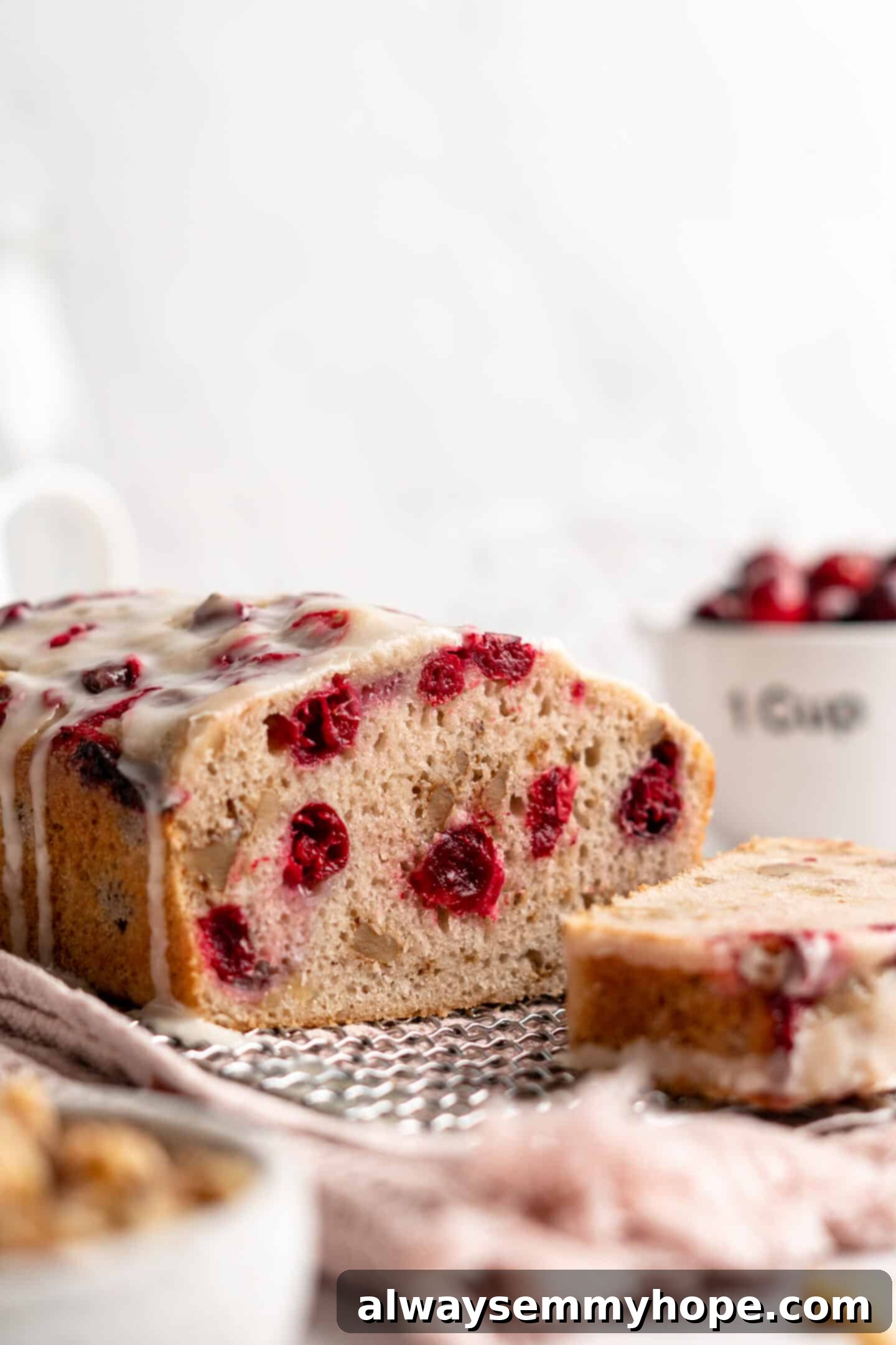 This moist cranberry walnut bread has just the right balance of sweetness, nuttiness, and tartness, and is a perfect holiday treat. A glistening loaf of cranberry orange bread, freshly glazed and sliced, rests on a cooling rack. A perfect slice is positioned in front, accompanied by a small bowl of fresh cranberries in the background, highlighting its deliciousness.