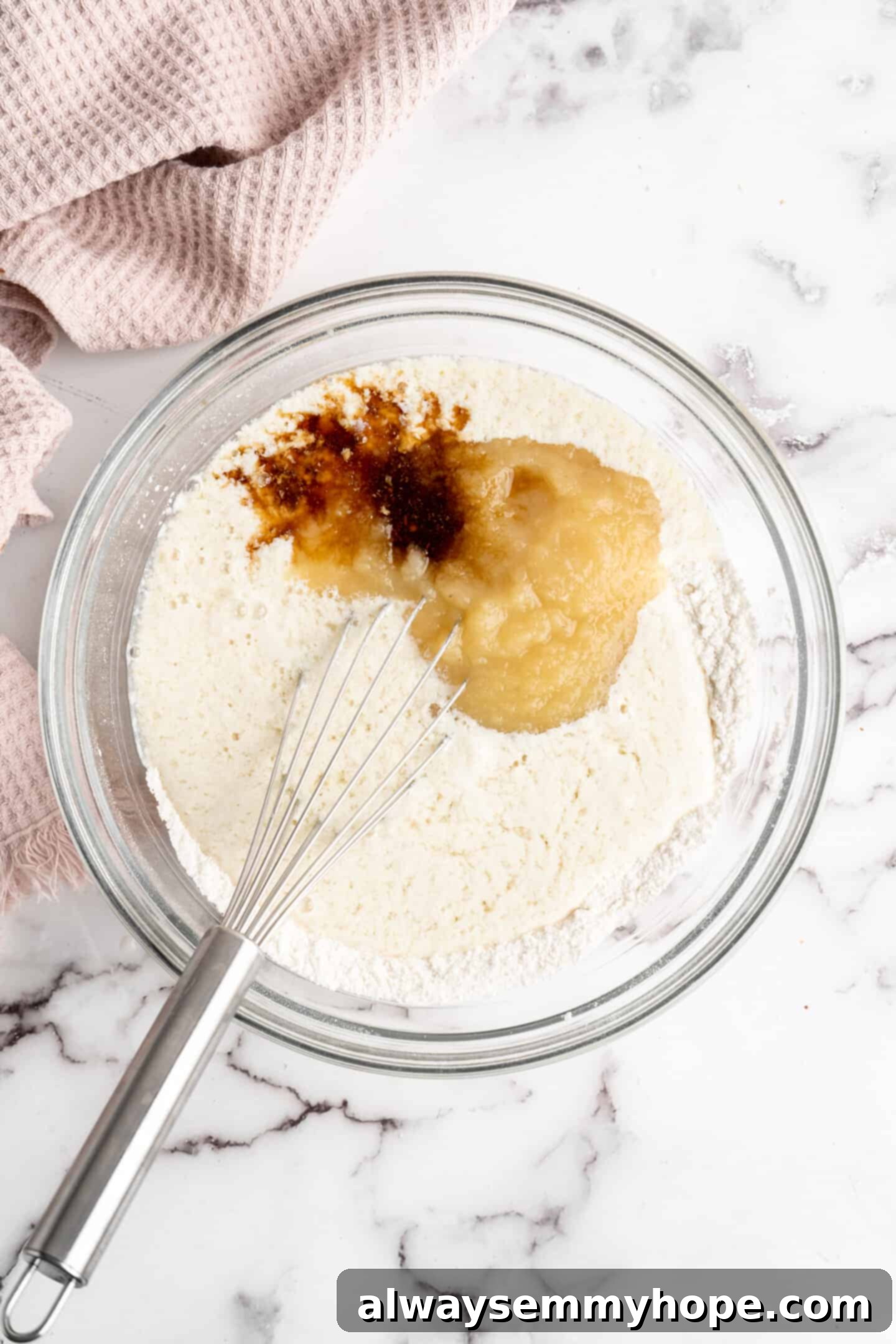 This moist cranberry walnut bread has just the right balance of sweetness, nuttiness, and tartness, and is a perfect holiday treat. A mixing bowl filled with the dry flour and sugar mixture, now topped with wet ingredients like applesauce, vegan buttermilk, and vanilla extract, with a whisk immersed, ready for mixing.