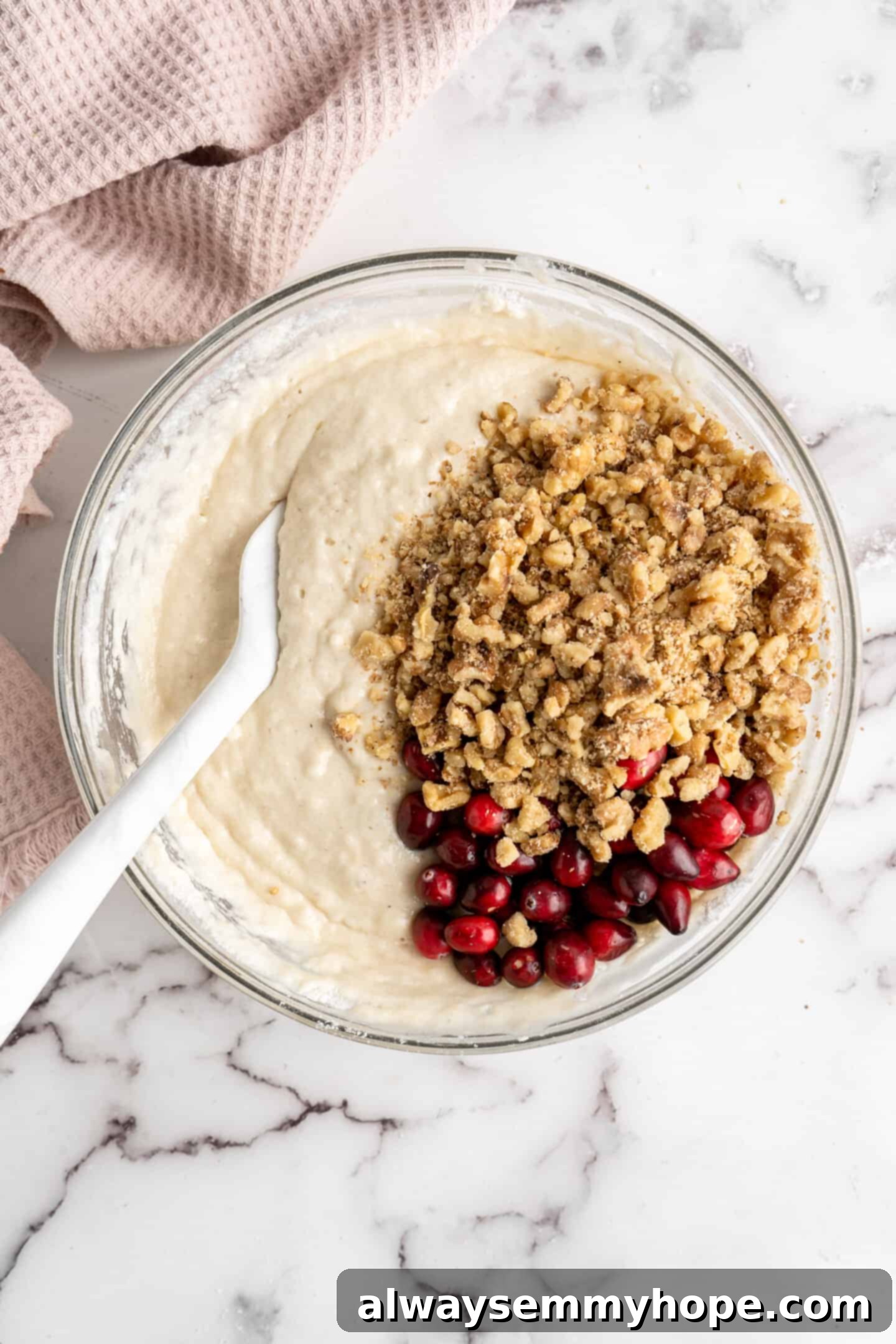 This moist cranberry walnut bread has just the right balance of sweetness, nuttiness, and tartness, and is a perfect holiday treat. A mixing bowl containing cranberry walnut bread batter, with chopped walnuts and flour-coated cranberries visibly sitting on top, ready to be folded in with a spatula.