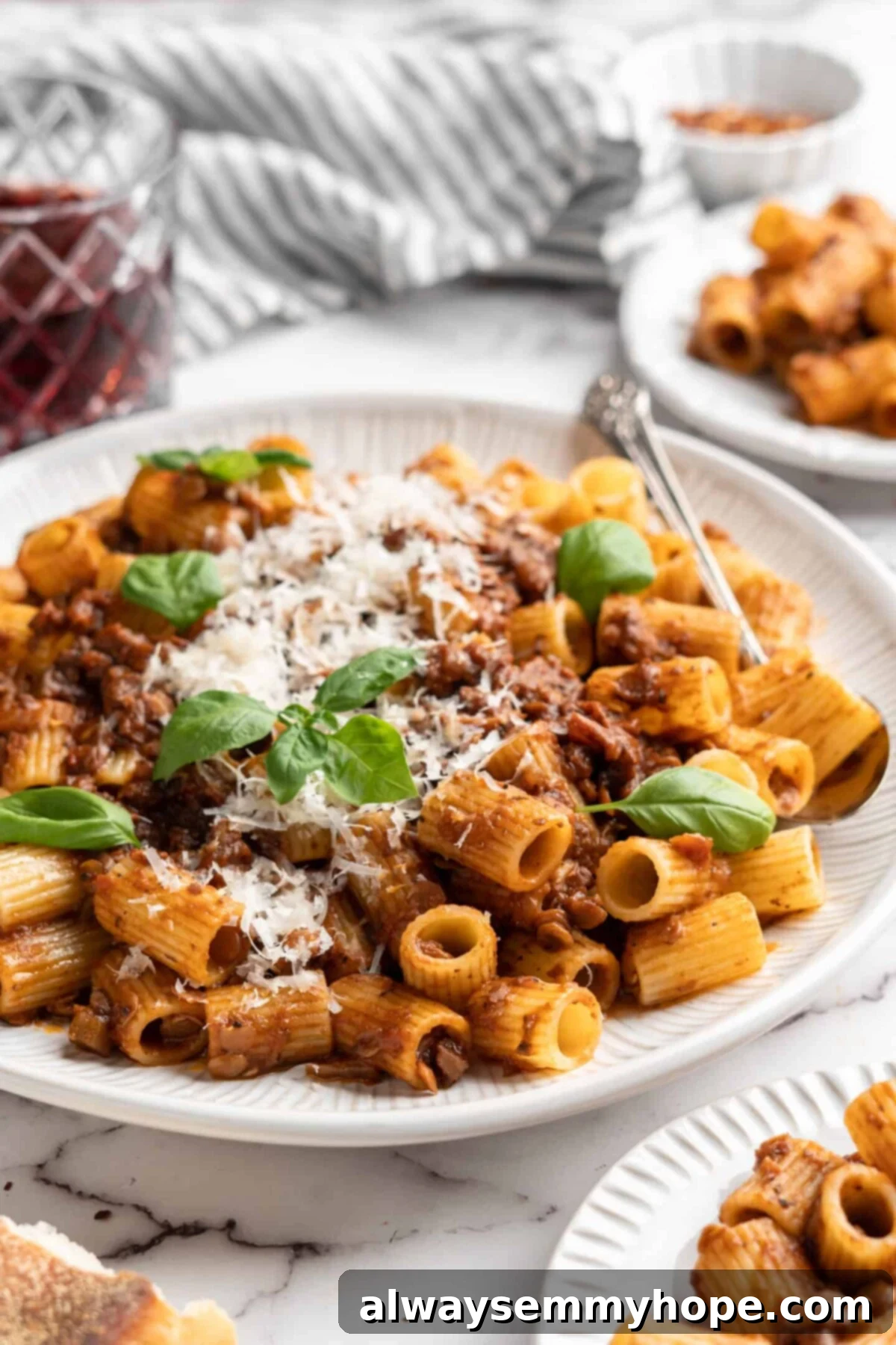 This easy walnut lentil bolognese recipe is a fun vegan take on a popular Italian pasta dish, and is full of tomato paste and veggies, perfect for sharing. A beautifully arranged plate of walnut lentil bolognese, artfully topped with vegan parmesan and fresh basil leaves, surrounded by additional plates of pasta, a rustic baguette, and a small bowl of chili flakes.