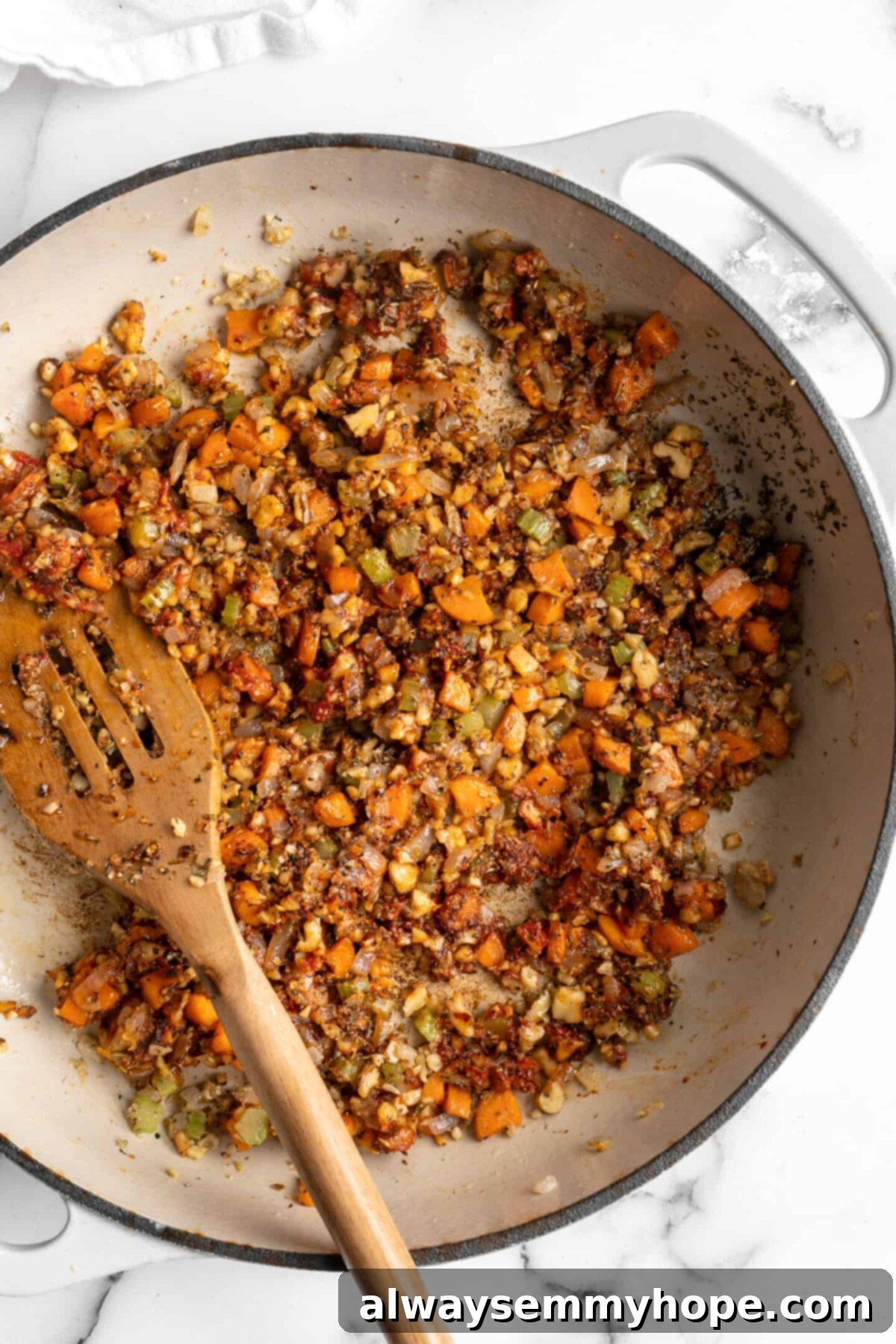 This easy walnut lentil bolognese recipe is a fun vegan take on a popular Italian pasta dish, and is full of tomato paste and veggies, illustrating the seasoned base. A pot with onions, carrots, celery, walnuts, garlic, tomato paste, and seasonings, with a wooden spoon in it, ready for the liquid ingredients.