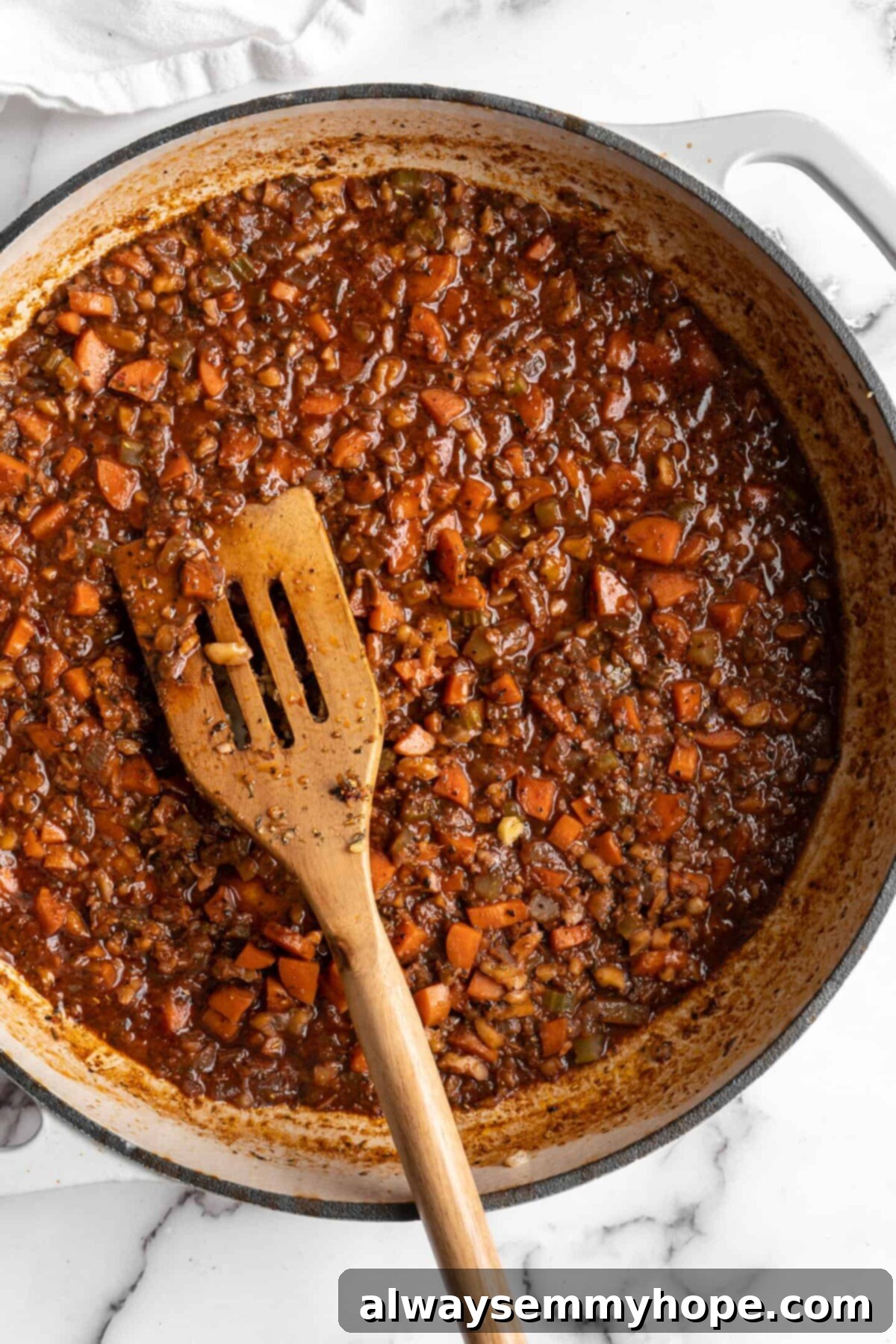 This easy walnut lentil bolognese recipe is a fun vegan take on a popular Italian pasta dish, and is full of tomato paste and veggies, showing the wine reduction. Red wine reducing with the sautéed veggies and walnuts for walnut lentil bolognese in a pot, with a wooden spoon, adding depth of flavor.
